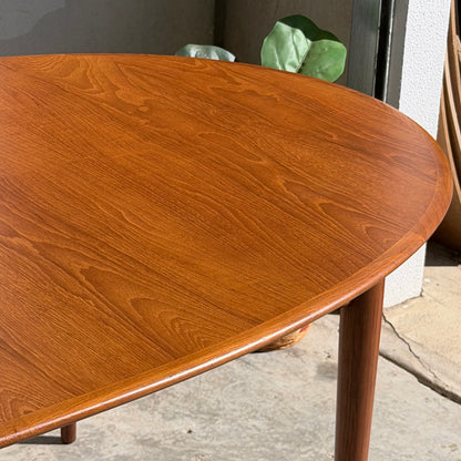 Sunlight highlights the Fully Restored Mid-Century Parker 91 Oval Teak Dining Suite with 6 Fler 64 Blackwood Chairs (Rediscover), its polished wood grain glowing on a concrete floor beside a potted plant, capturing classic Australian mid-century style.