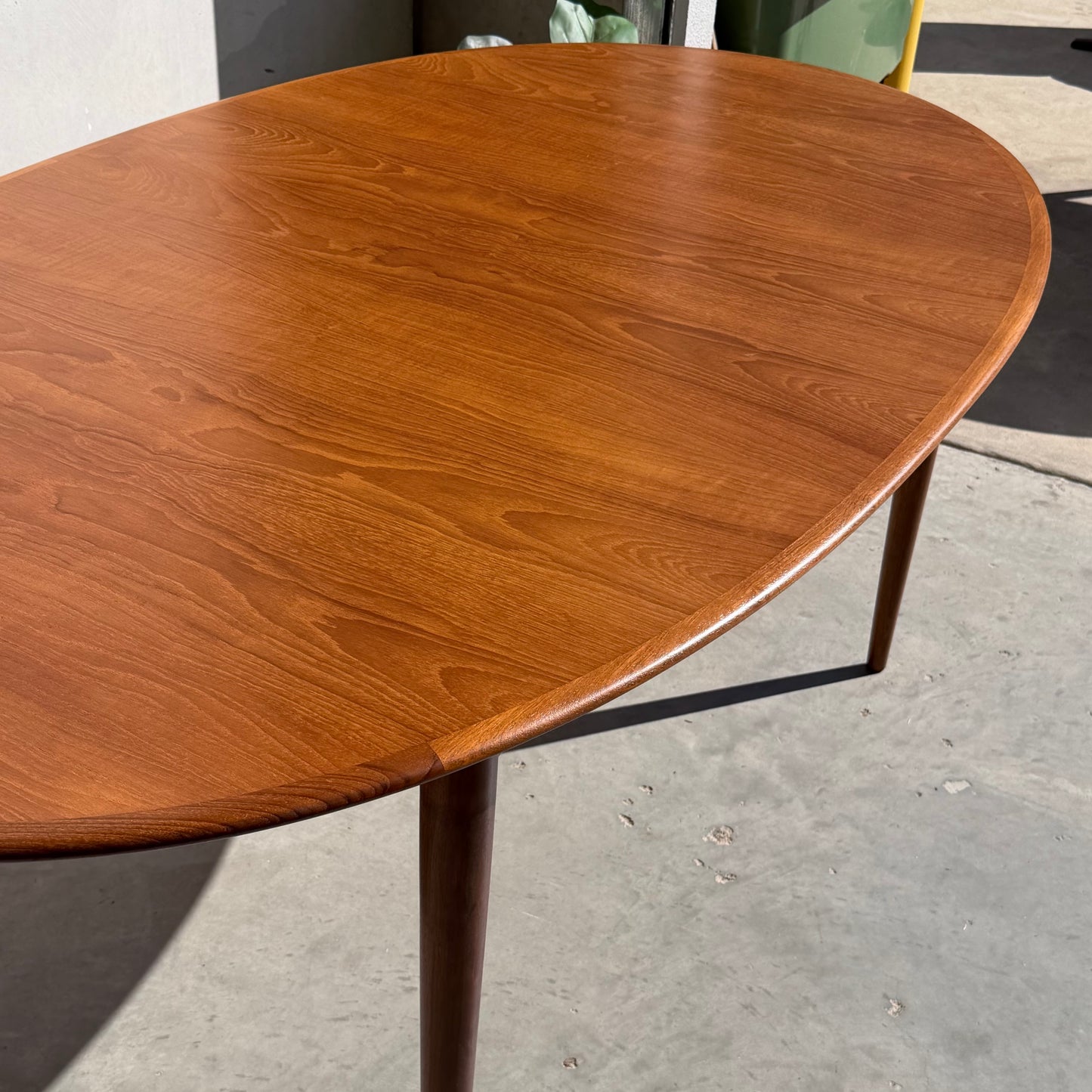 A fully restored Mid-Century Parker 91 oval teak dining suite with 6 Fler 64 blackwood chairs (Rediscover) sits on concrete in sunlight, its rich wood grain and warm brown finish reflecting classic Australian style.
