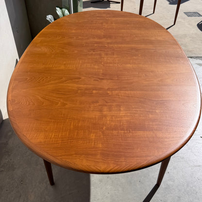 A fully restored Parker No.91 Fixed Top Oval Table (Rediscover) with a smooth, polished surface sits on a concrete floor near a wall, sunlight casting soft shadows across its teak finish.