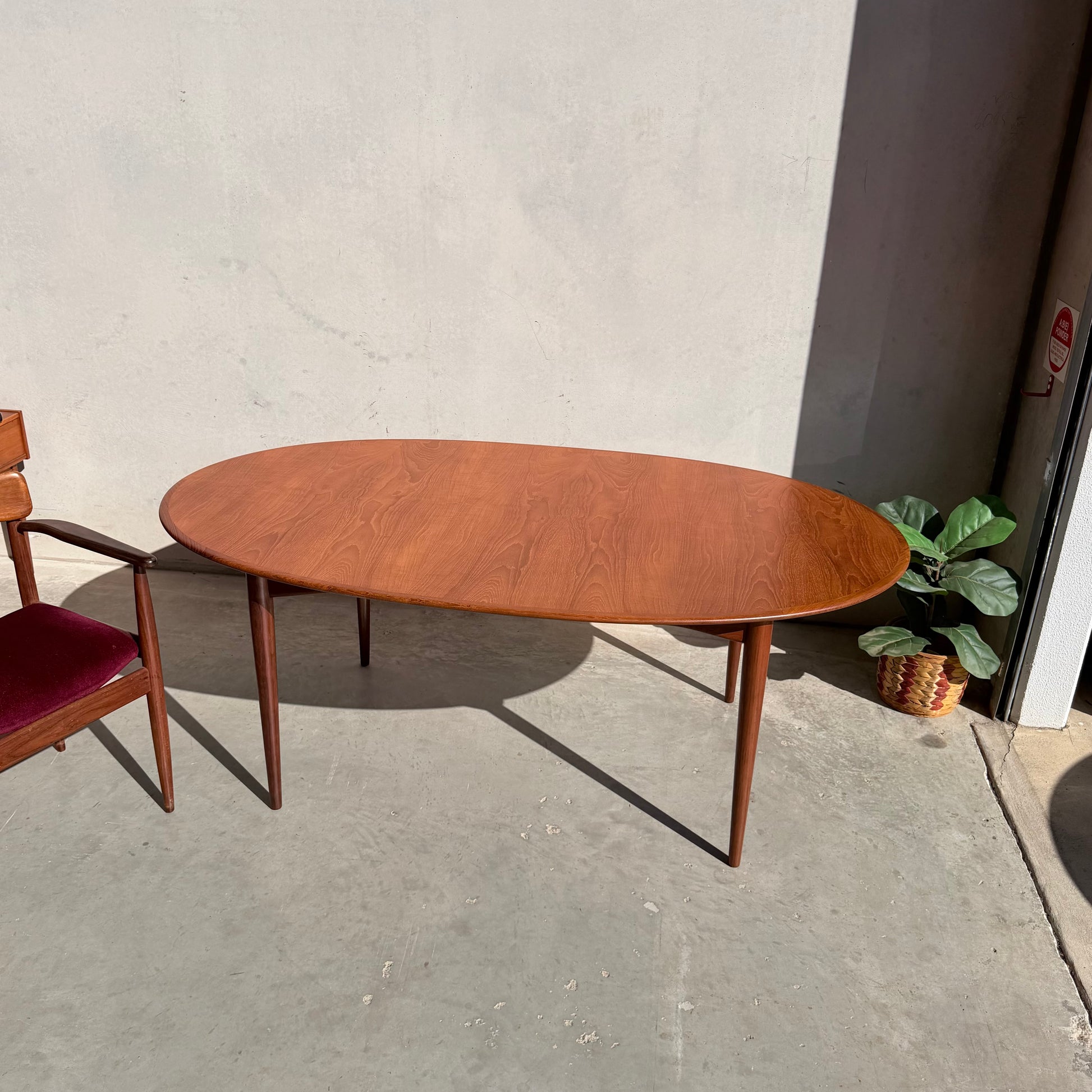 A fully restored Parker No.91 Fixed Top Oval Table (Rediscover) in mid-century modern style stands on a concrete floor near a wooden chair with a red seat, a potted plant, and a sunlit bare wall.
