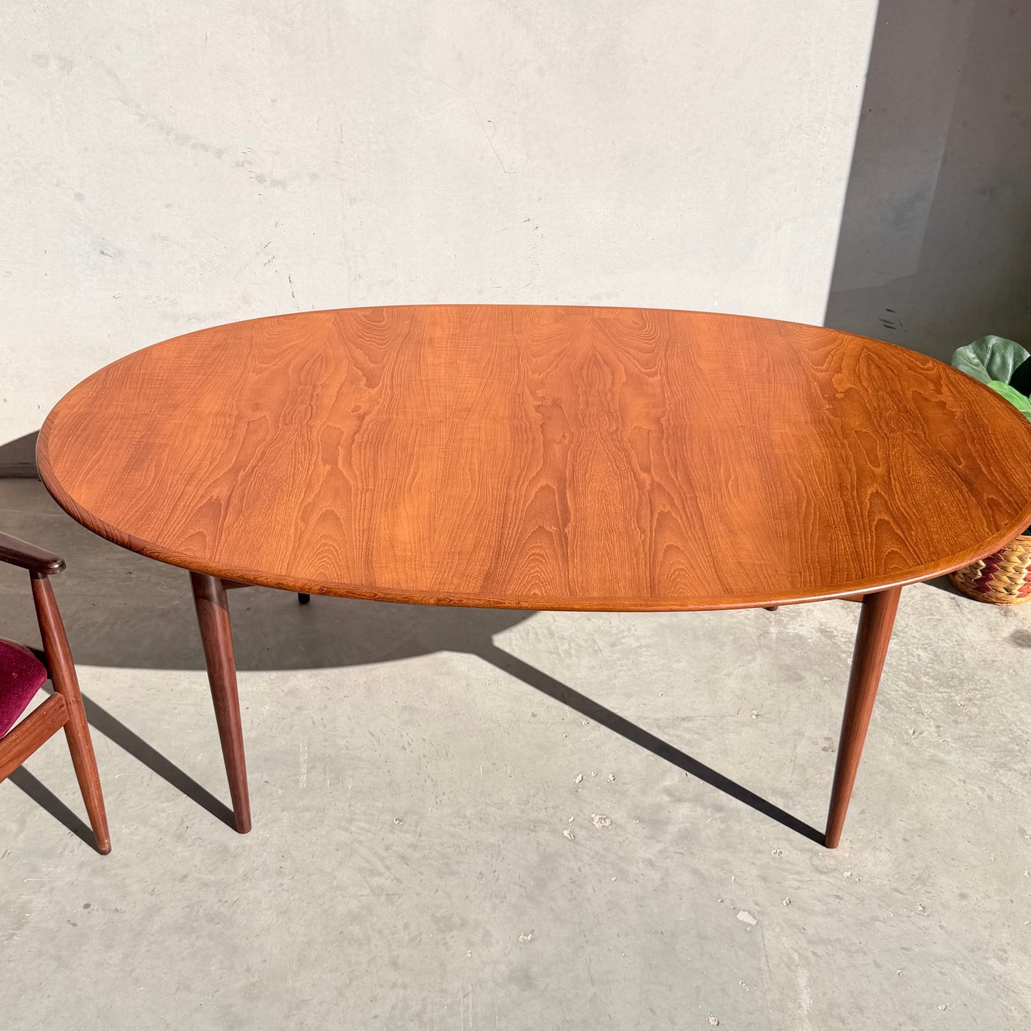 A Fully Restored Parker No.91 Fixed Top Oval Table (Rediscover) with tapered legs stands on a light concrete floor. Part of a chair with a red cushion appears on the left as sunlight creates soft shadows on the wall and floor.