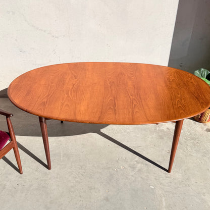 A Fully Restored Parker No.91 Fixed Top Oval Table (Rediscover) with tapered legs stands on a light concrete floor. Part of a chair with a red cushion appears on the left as sunlight creates soft shadows on the wall and floor.