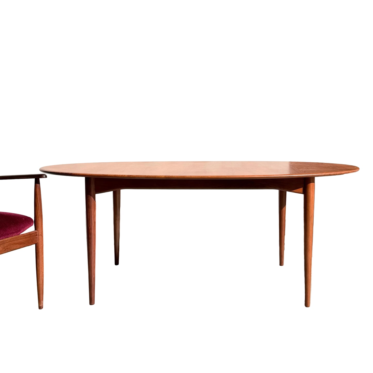 A Fully Restored Parker No.91 Fixed Top Oval Table with tapered legs stands on a concrete floor beside a wooden chair with a red seat; nearby, a potted plant rests against a plain, sunlit wall.
