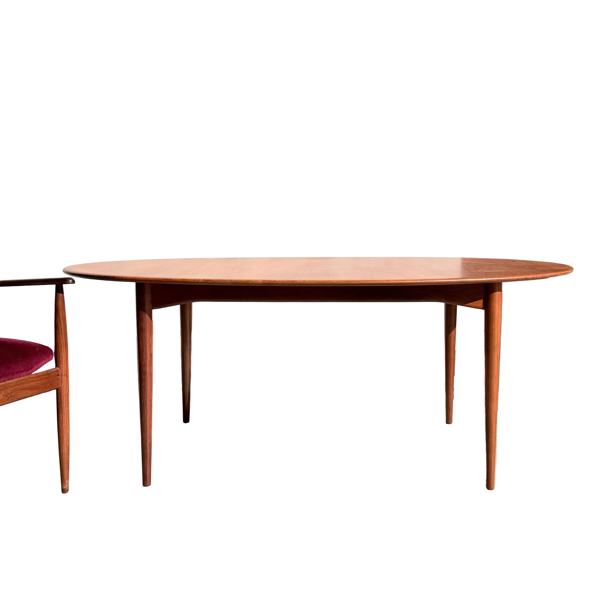 A Fully Restored Parker No.91 Fixed Top Oval Table with tapered legs stands on a concrete floor beside a wooden chair with a red seat; nearby, a potted plant rests against a plain, sunlit wall.