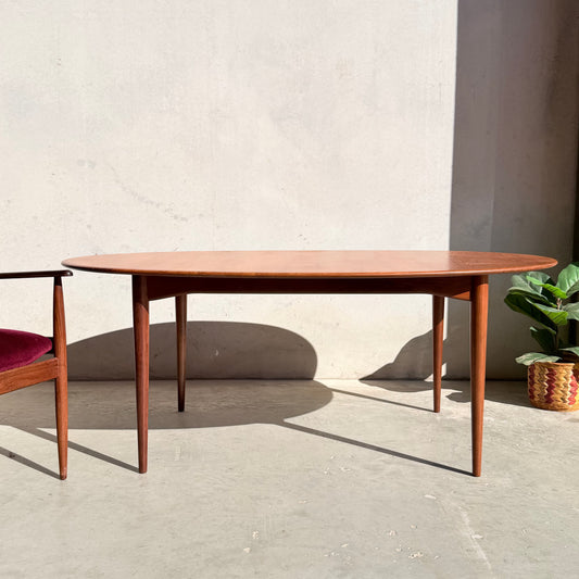 A Fully Restored Parker No.91 Fixed Top Oval Table with tapered legs stands on a concrete floor beside a wooden chair with a red seat; nearby, a potted plant rests against a plain, sunlit wall.