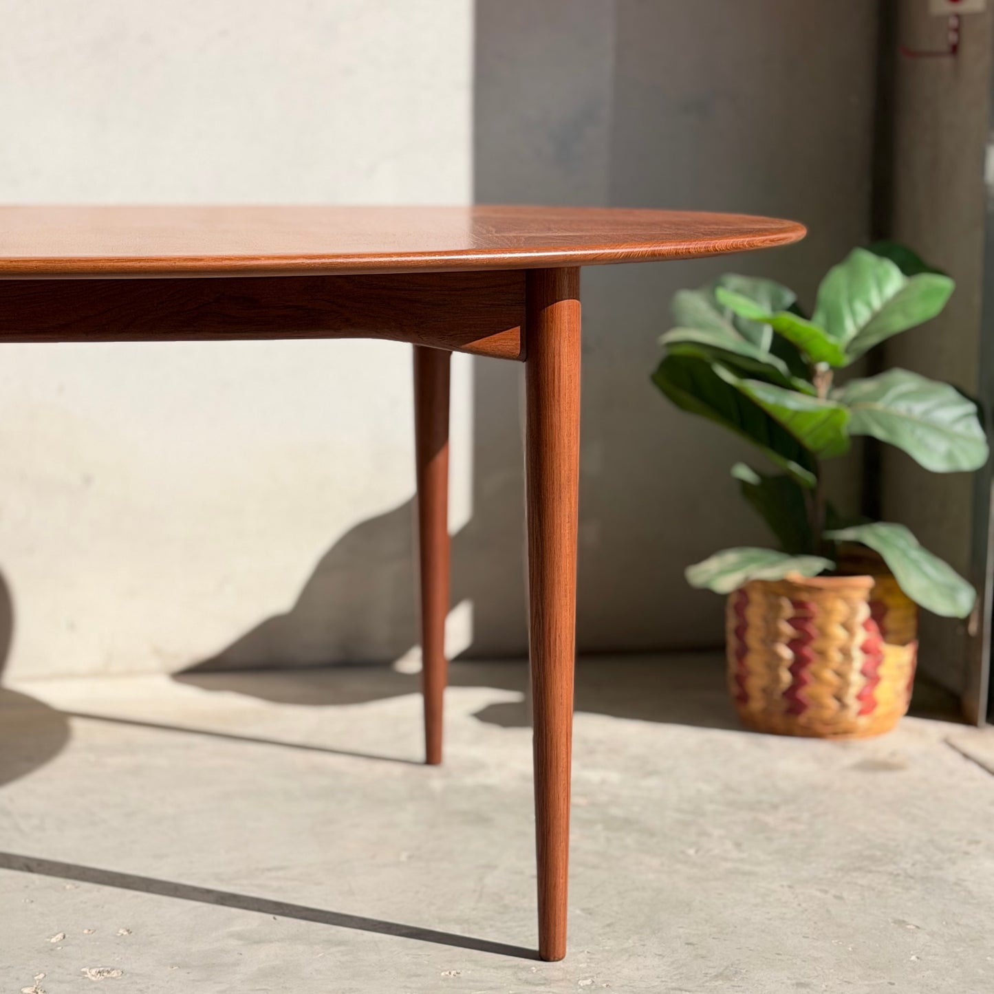 The Fully Restored Parker No.91 Fixed Top Oval Table (Rediscover) with tapered legs stands on a sunlit cement floor beside a green potted plant in a woven basket, sunlight casting defined shadows across the floor and wall.