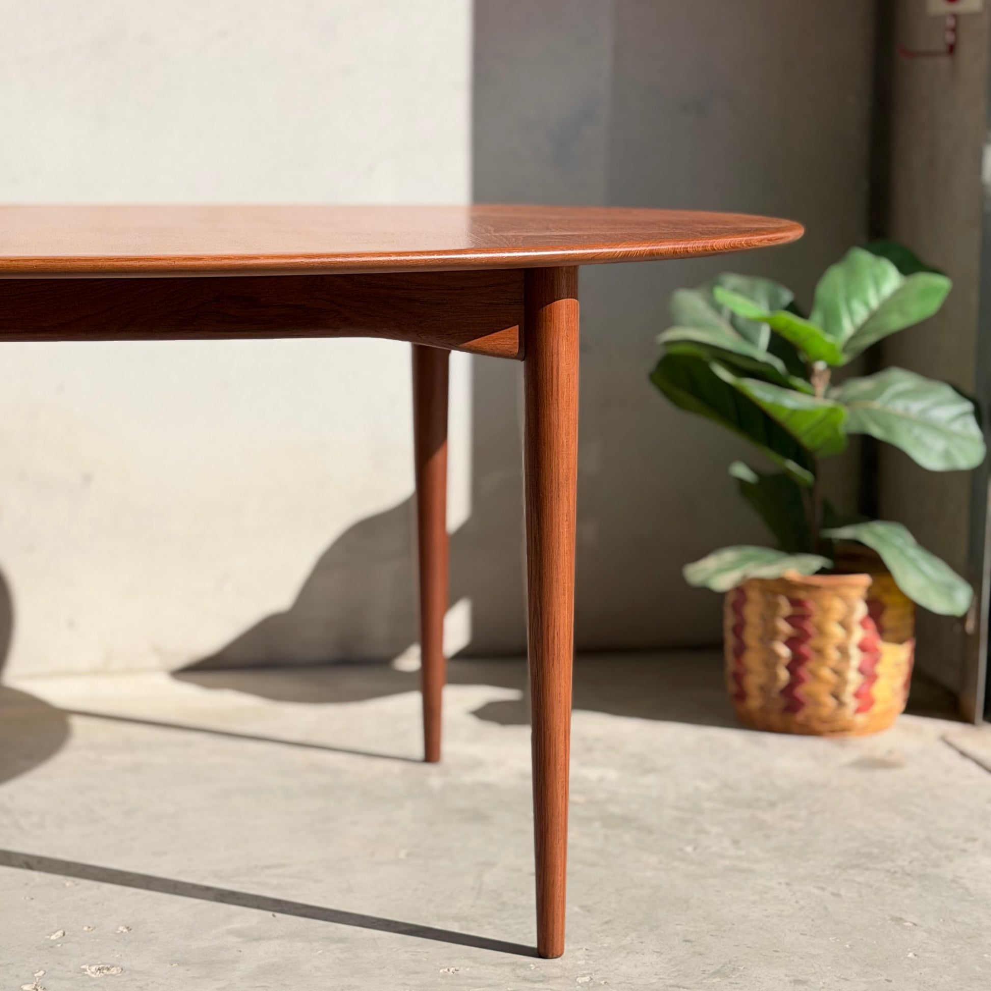 The Fully Restored Parker No.91 Fixed Top Oval Table (Rediscover) with tapered legs stands on a sunlit cement floor beside a green potted plant in a woven basket, sunlight casting defined shadows across the floor and wall.