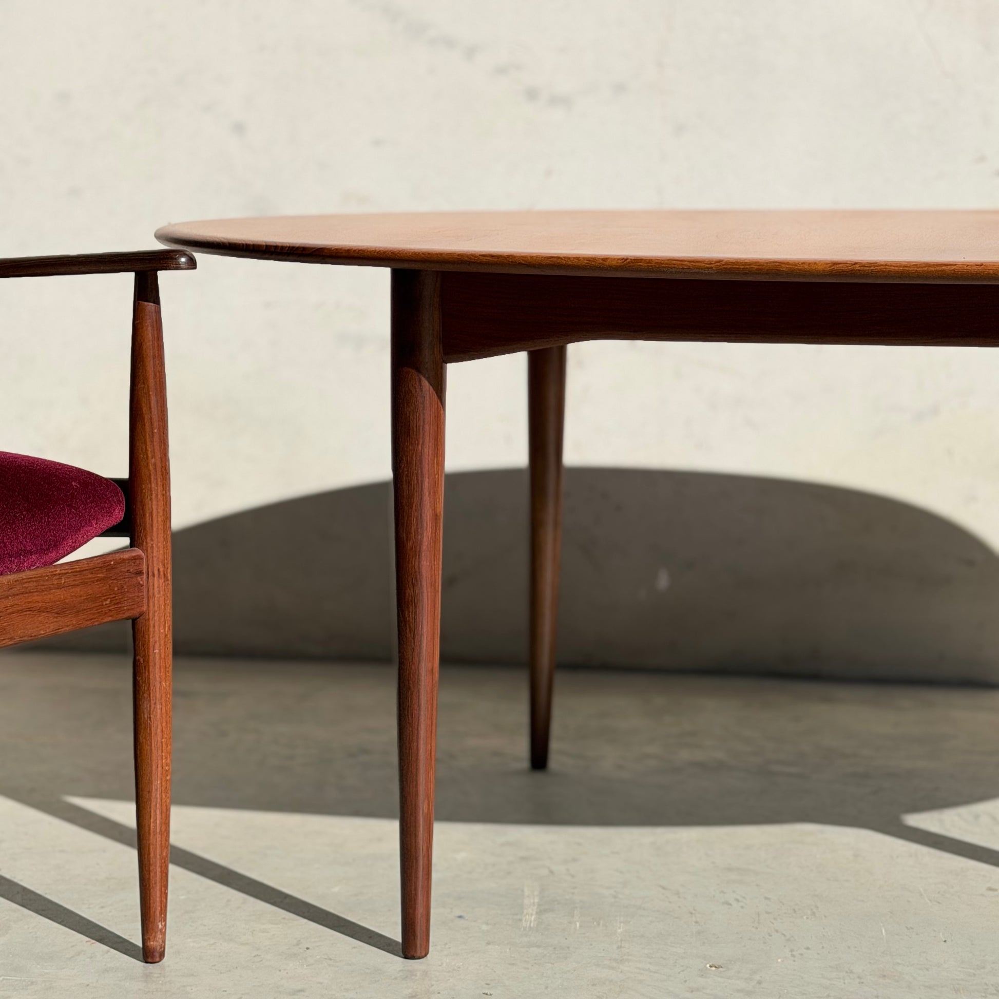 A Fully Restored Parker No.91 Fixed Top Oval Table (Rediscover) is paired with a matching wooden chair with a red upholstered seat, all set against a light-colored wall in bright sunlight.