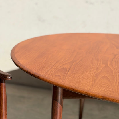 A close-up of a Fully Restored Parker No.91 Fixed Top Oval Table (Rediscover) showcases its grain patterns, paired with part of a matching wooden chair, set against a plain, light-colored background.