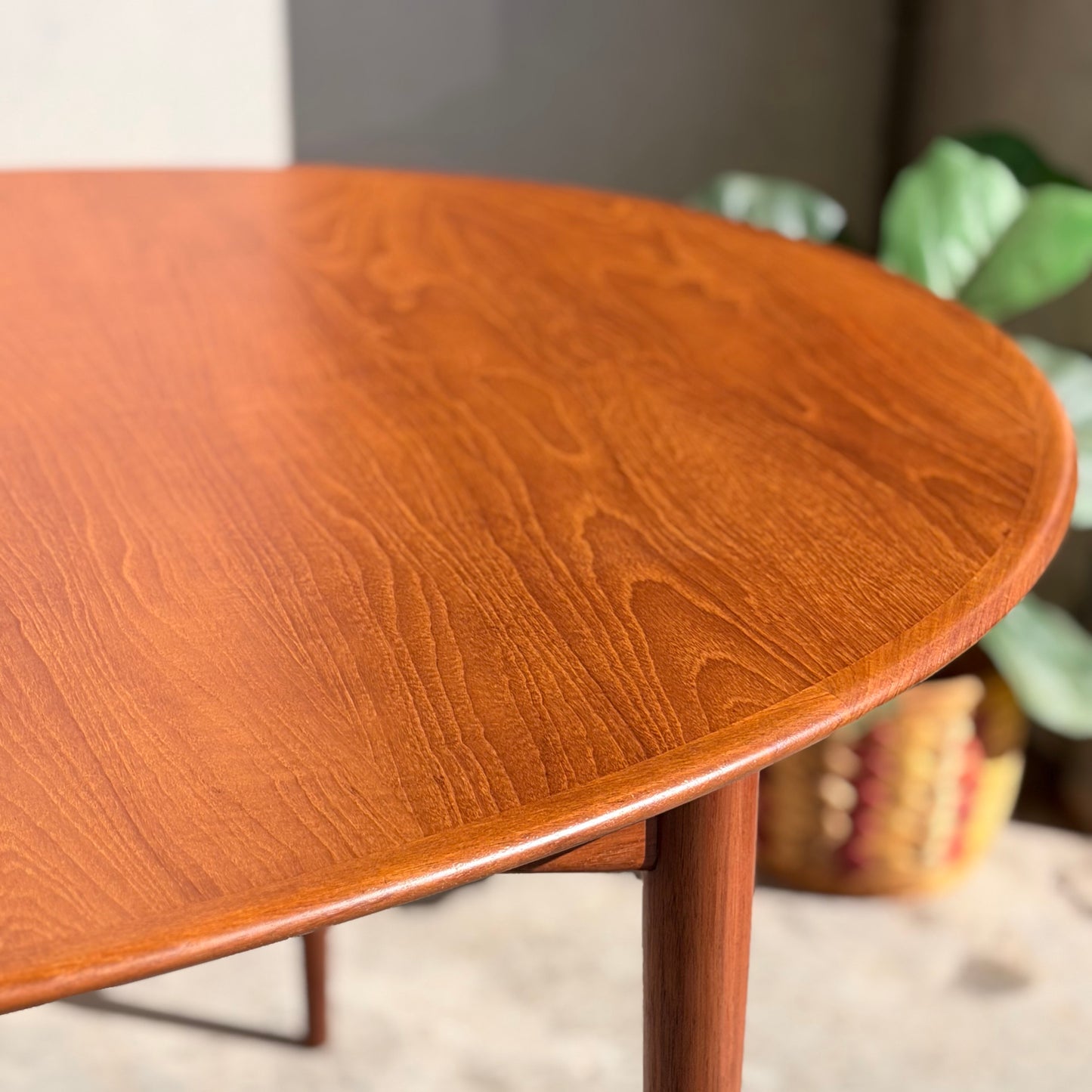 Close-up of the Fully Restored Parker No.91 Fixed Top Oval Table (Rediscover), showcasing its smooth, polished teak surface and visible grain. A blurred green plant and woven basket appear in the background.