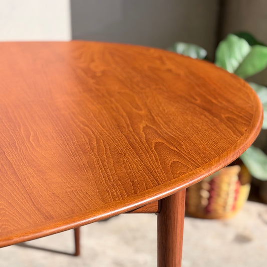 Close-up of the Fully Restored Parker No.91 Fixed Top Oval Table (Rediscover), showcasing its smooth, polished teak surface and visible grain. A blurred green plant and woven basket appear in the background.