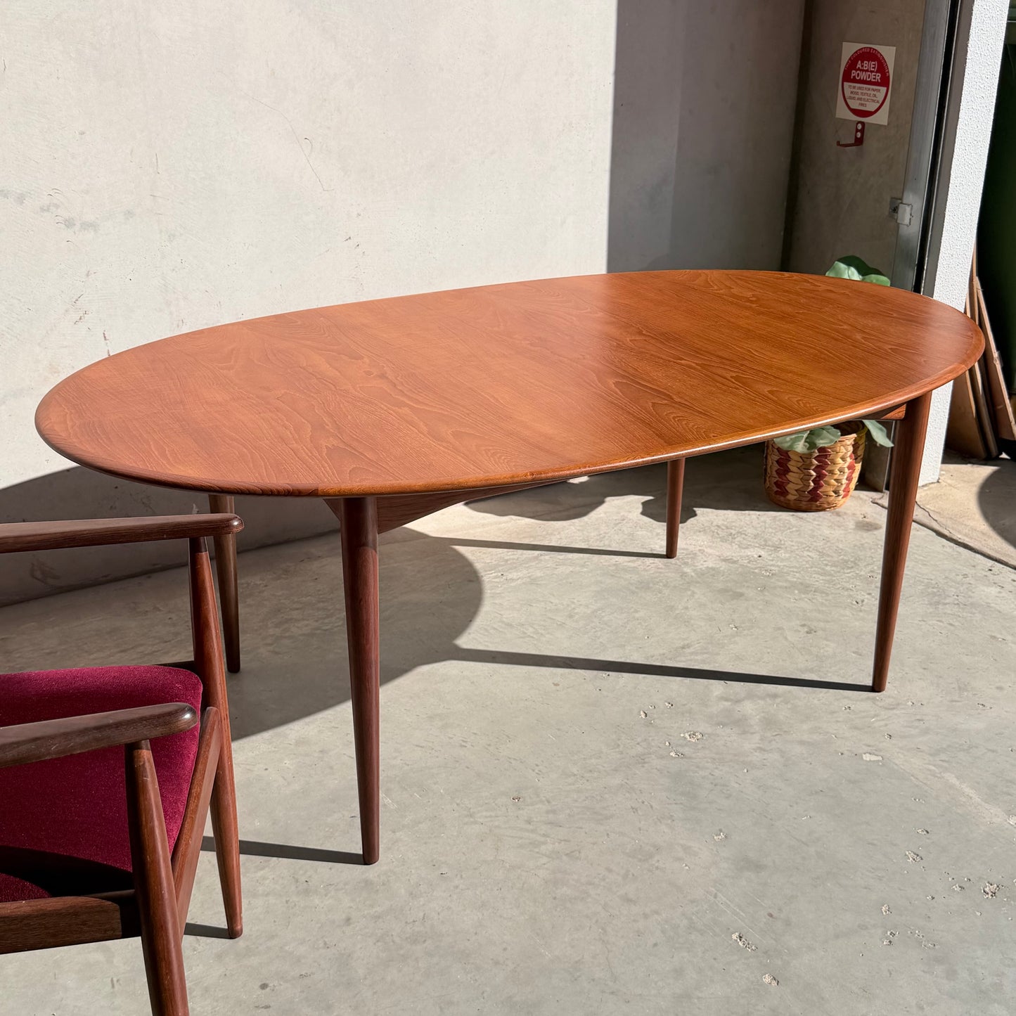 A Fully Restored Parker No.91 Fixed Top Oval Table (Rediscover) sits on a concrete floor near a wall, sunlight casting gentle shadows. In the lower left corner, part of a wooden chair with a red seat cushion is visible.