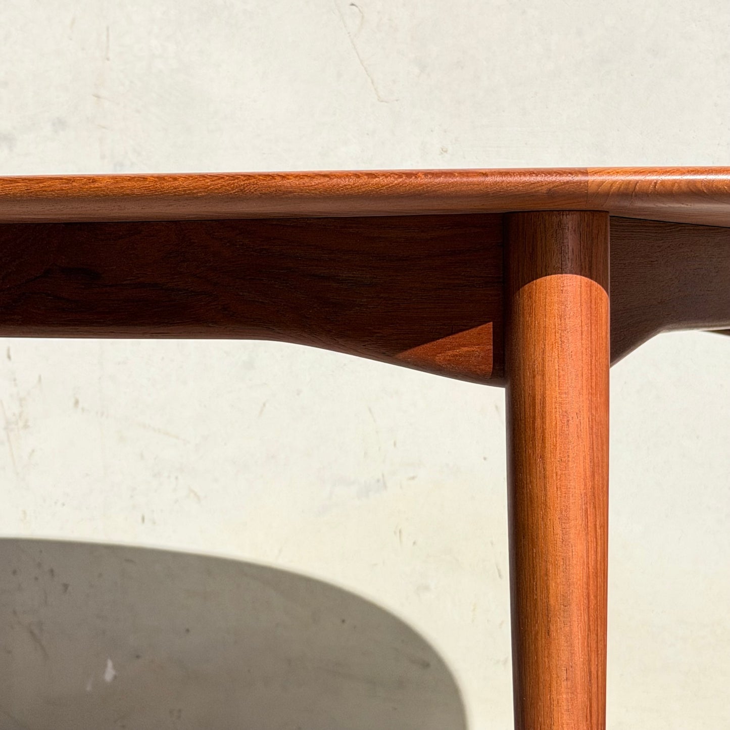 Close-up of the Fully Restored Parker No.91 Fixed Top Oval Table (Rediscover) corner with smooth, rounded edges and a cylindrical leg, sunlight casting shadows on its teak surface against a light wall.