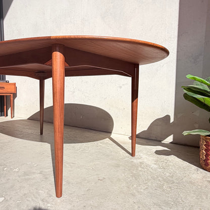 A Fully Restored Parker No.91 Fixed Top Oval Table (Rediscover) with tapered legs stands on a sunlit concrete floor by a potted plant, its gleaming teak surface contrasting against a light-colored wall.