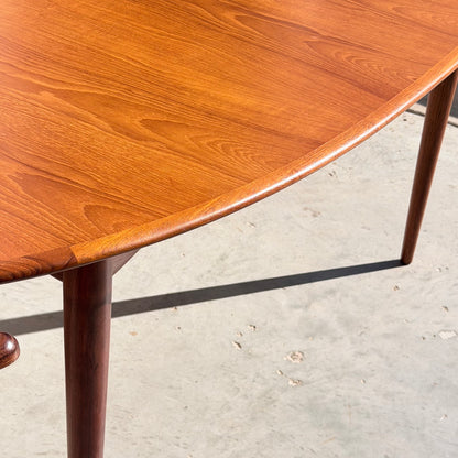 Close-up of the Fully Restored Parker No.91 Fixed Top Oval Table (Rediscover) shows its smooth, polished teak surface and rounded edge, highlighting the wood grain and three slender tapered legs on a light concrete floor.