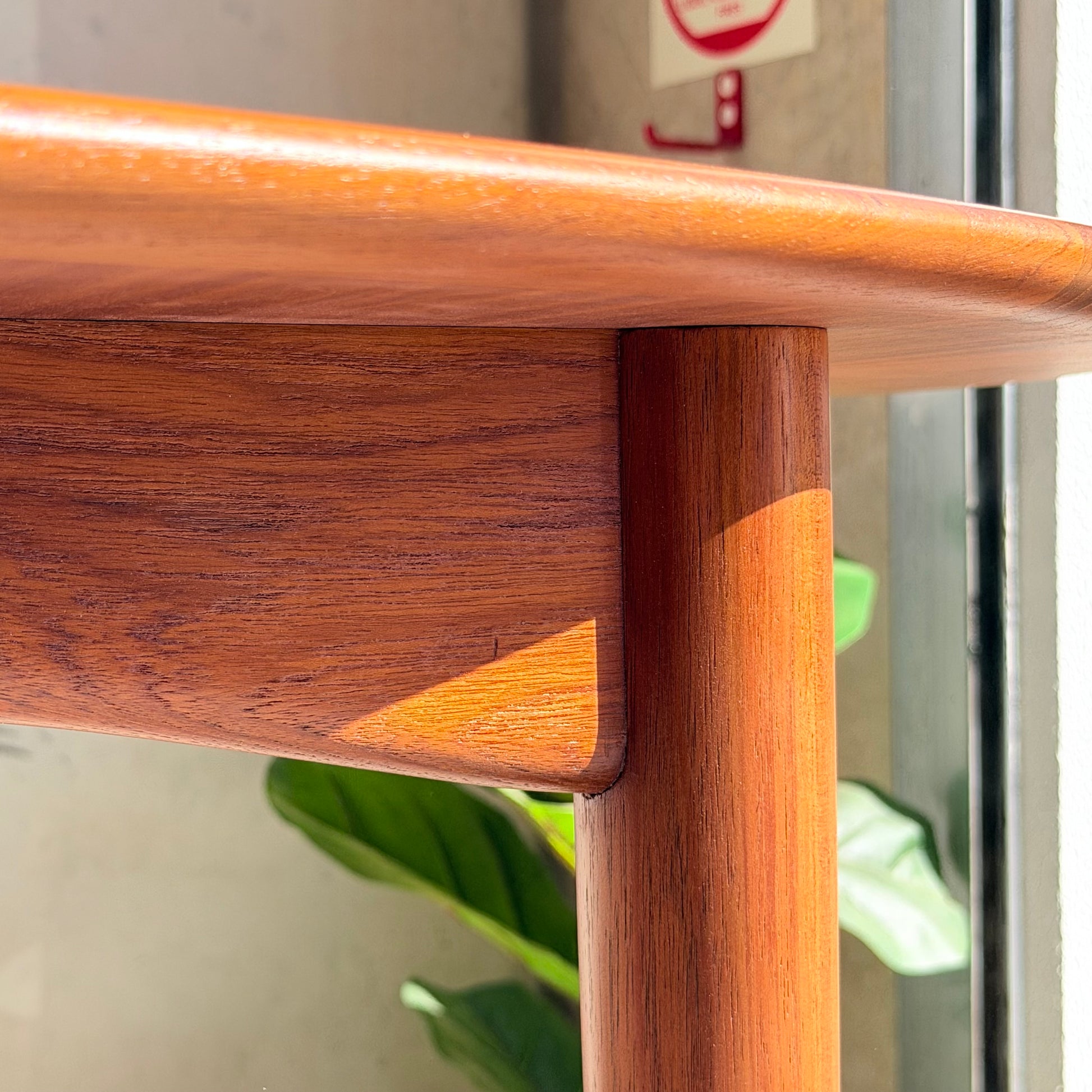 Close-up of the Fully Restored Parker No.91 Fixed Top Oval Table (Rediscover) corner with smooth, rounded teak edges and rich wood grain; sunlight highlights the finish while a blurred green plant and wall with fire alarm appear in the background.
