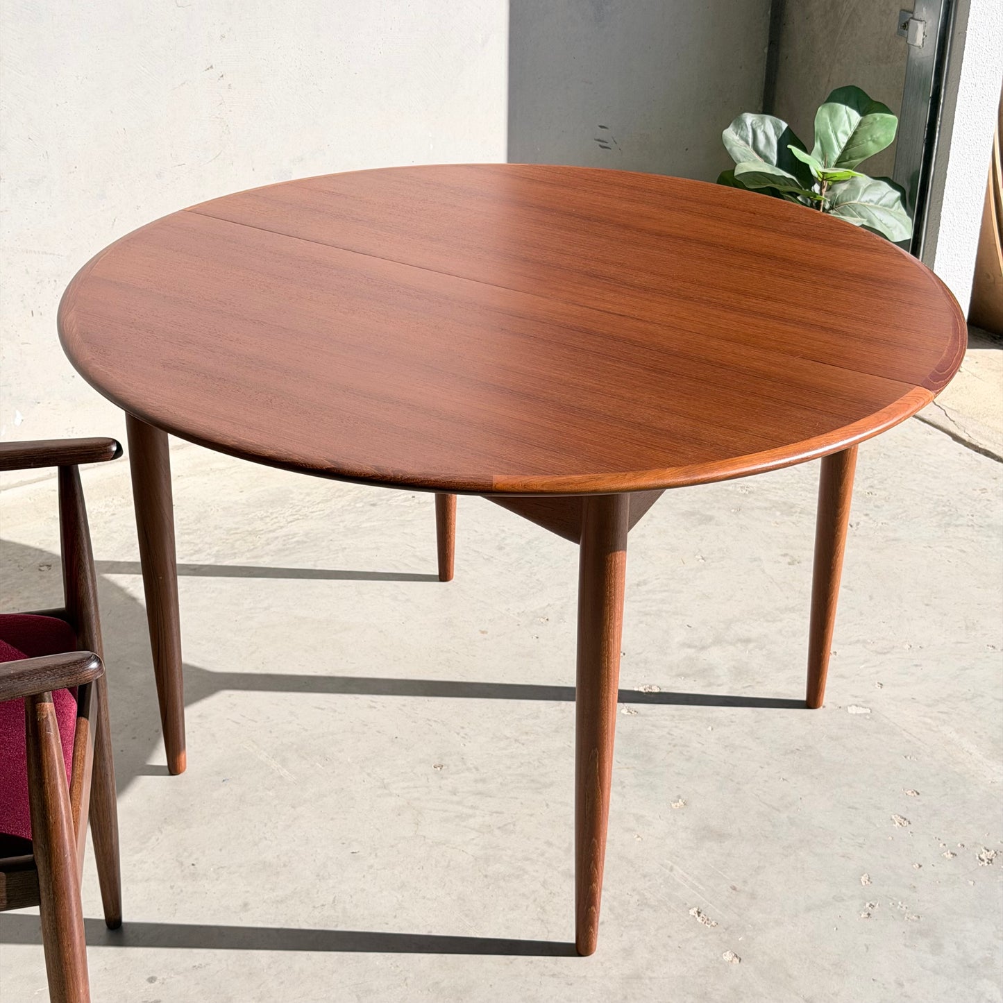 A Fully Restored Parker 90 Round Extension Butterfly Table stands on a light concrete floor with sunlight streaming in from the right, complemented by a matching chair with a red cushion and a green potted plant in the background.