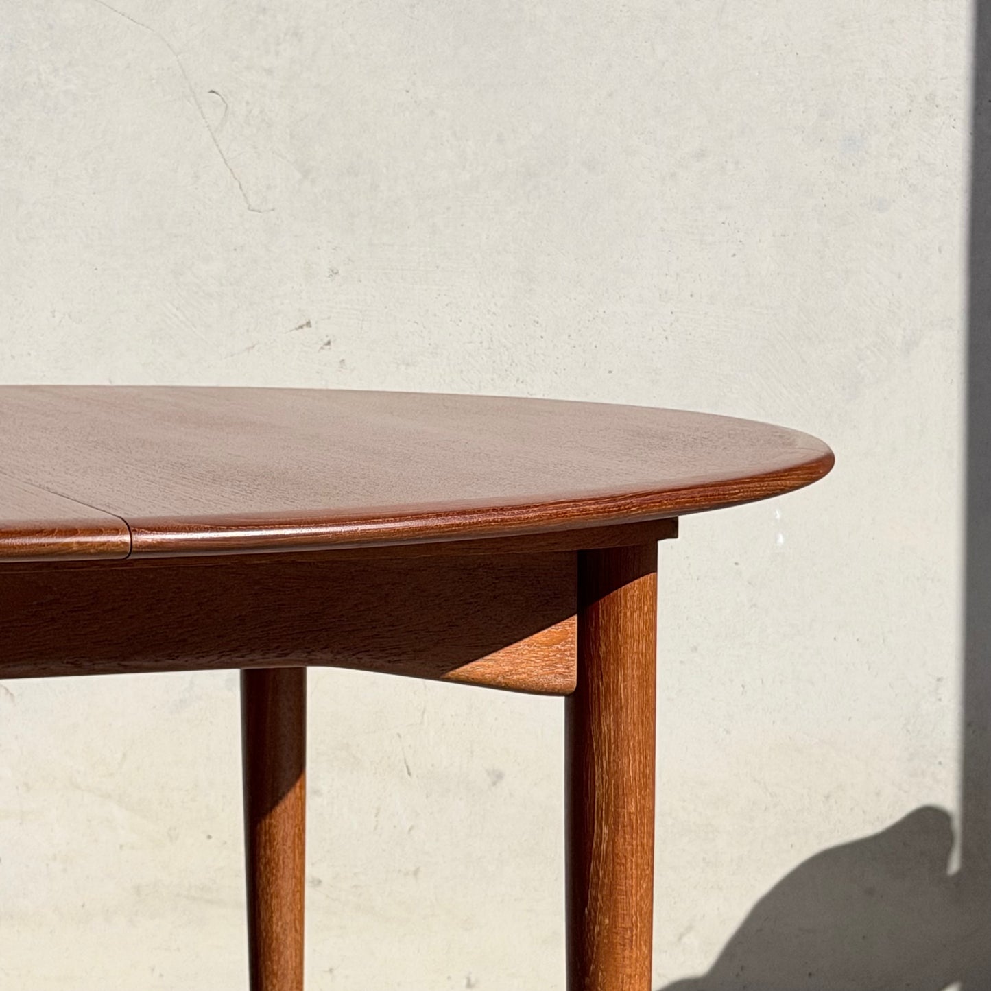 A close-up of the Fully Restored Parker 90 Round Extension Butterfly Table Only (Rediscover) with rounded edges and cylindrical legs, set against a light wall as sunlight casts shadows on the surface.