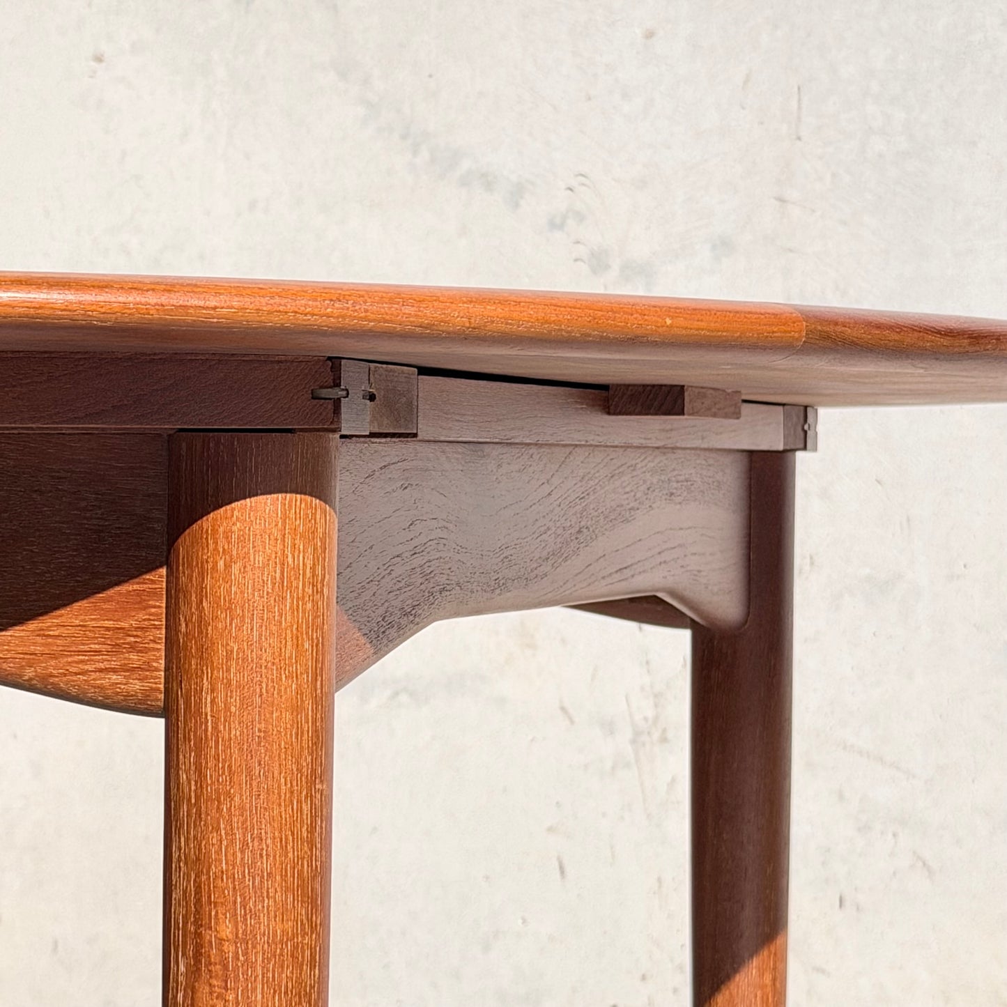 Close-up of a Fully Restored Parker 90 Round Extension Butterfly Table Only (Rediscover) with rounded edges and cylindrical legs, set against a light wall. Sunlight highlights its mid-century modern design and smooth teak finish.