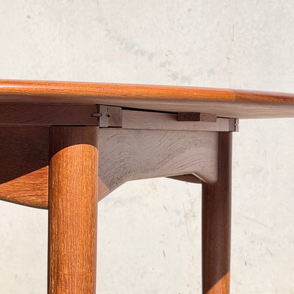 Close-up of a Fully Restored Parker 90 Round Extension Butterfly Table Only (Rediscover) with rounded edges and cylindrical legs, set against a light wall. Sunlight highlights its mid-century modern design and smooth teak finish.