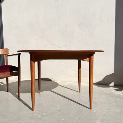 A Fully Restored Parker 90 Round Extension Butterfly Table stands on a light floor beside a wooden chair with a red seat cushion. Sunlight creates sharp shadows, set against a plain, light-colored wall.