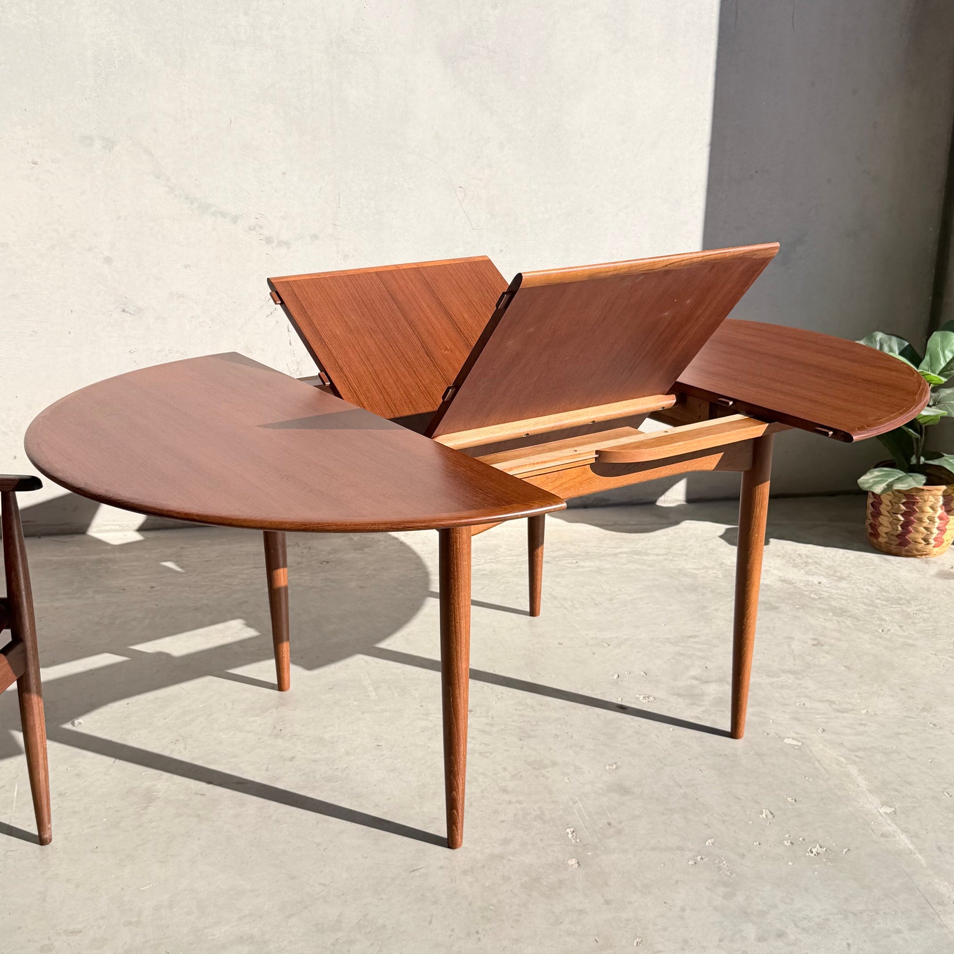 A fully restored Parker 90 Round Extension Butterfly Table (Rediscover) stands with its extension leaves partially open on a concrete floor near a wall and a potted plant.