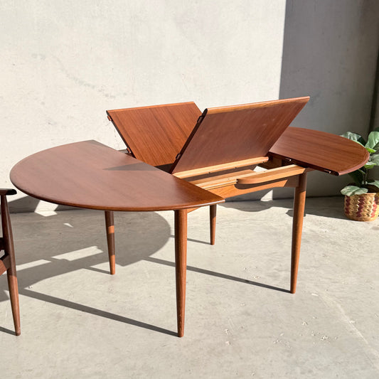 A fully restored Parker 90 Round Extension Butterfly Table (Rediscover) stands with its extension leaves partially open on a concrete floor near a wall and a potted plant.
