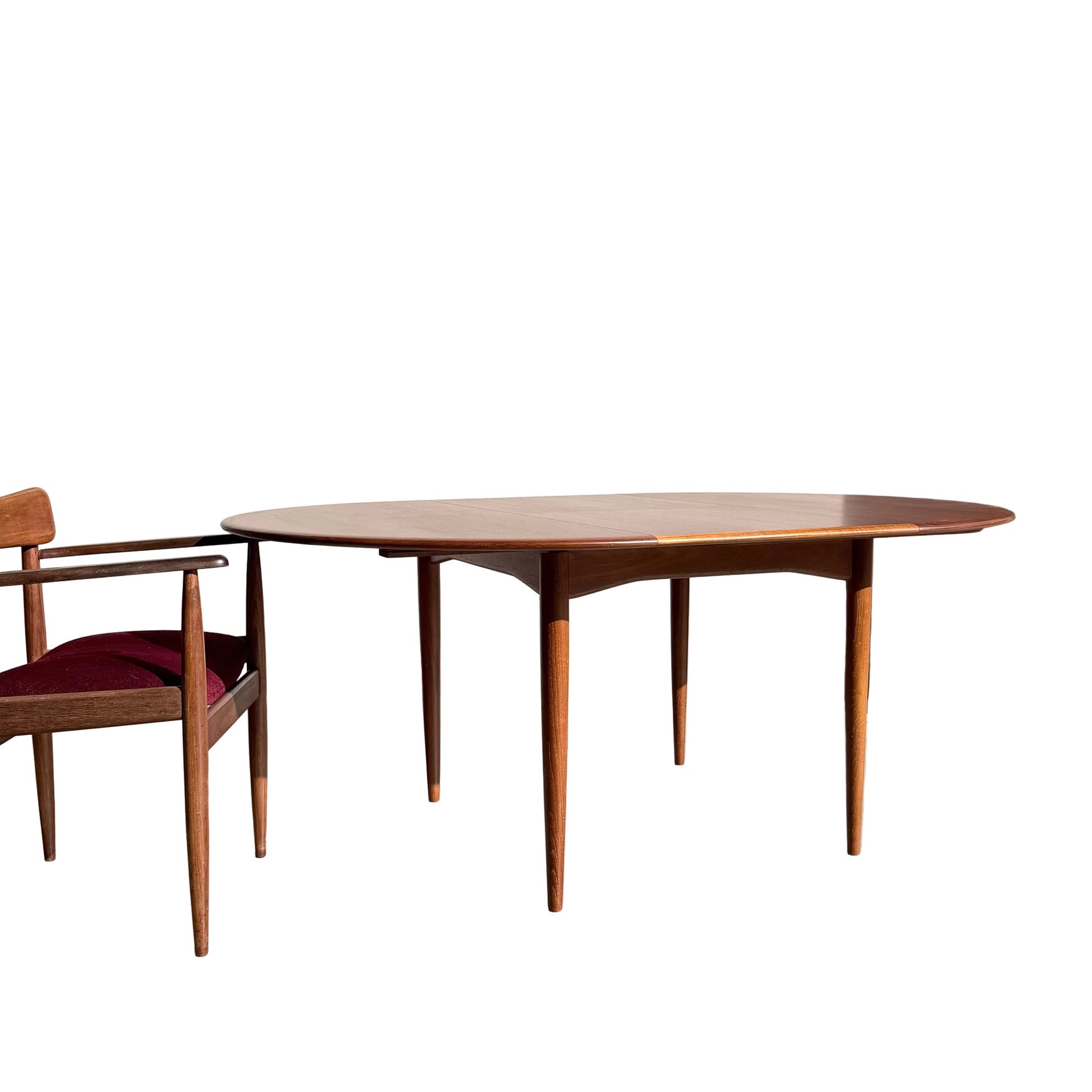 A Fully Restored Parker 90 Round Extension Butterfly Table (Rediscover) with tapered legs is set on a sunlit concrete floor near a matching chair with a burgundy cushion. A small potted plant in a woven basket rests by the light-colored wall.