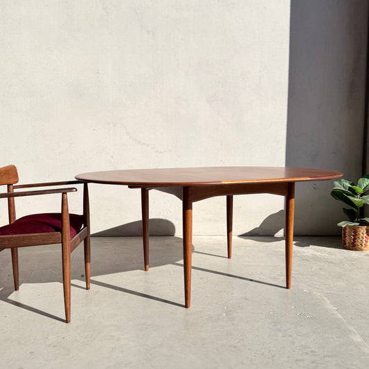 A Fully Restored Parker 90 Round Extension Butterfly Table (Rediscover) with tapered legs is set on a sunlit concrete floor near a matching chair with a burgundy cushion. A small potted plant in a woven basket rests by the light-colored wall.