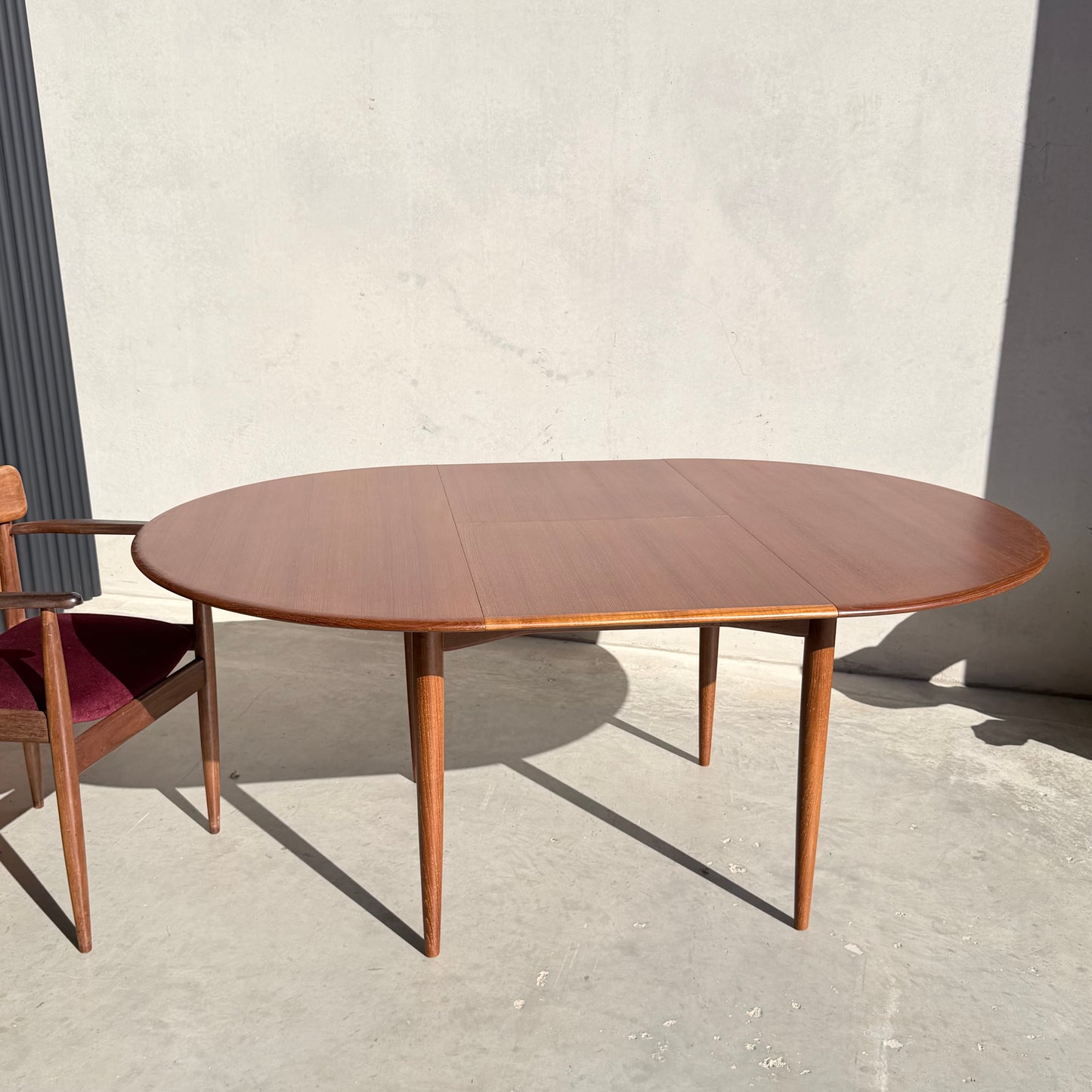 A Fully Restored Parker 90 Round Extension Butterfly Table (Rediscover) with tapered legs stands on a sunlit concrete floor next to a wooden chair with a red seat cushion.