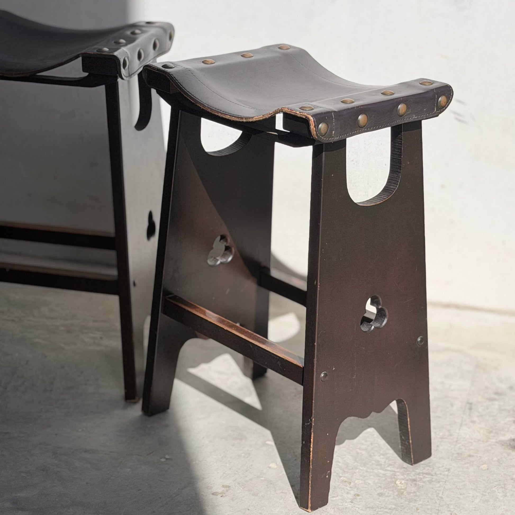 A 1970s Australian Mid-Century Modern Spanish Revival Davis stool with a brown leather seat and metal studs sits on a sunlit concrete floor beside a matching stool; part of the Revive set of 3.