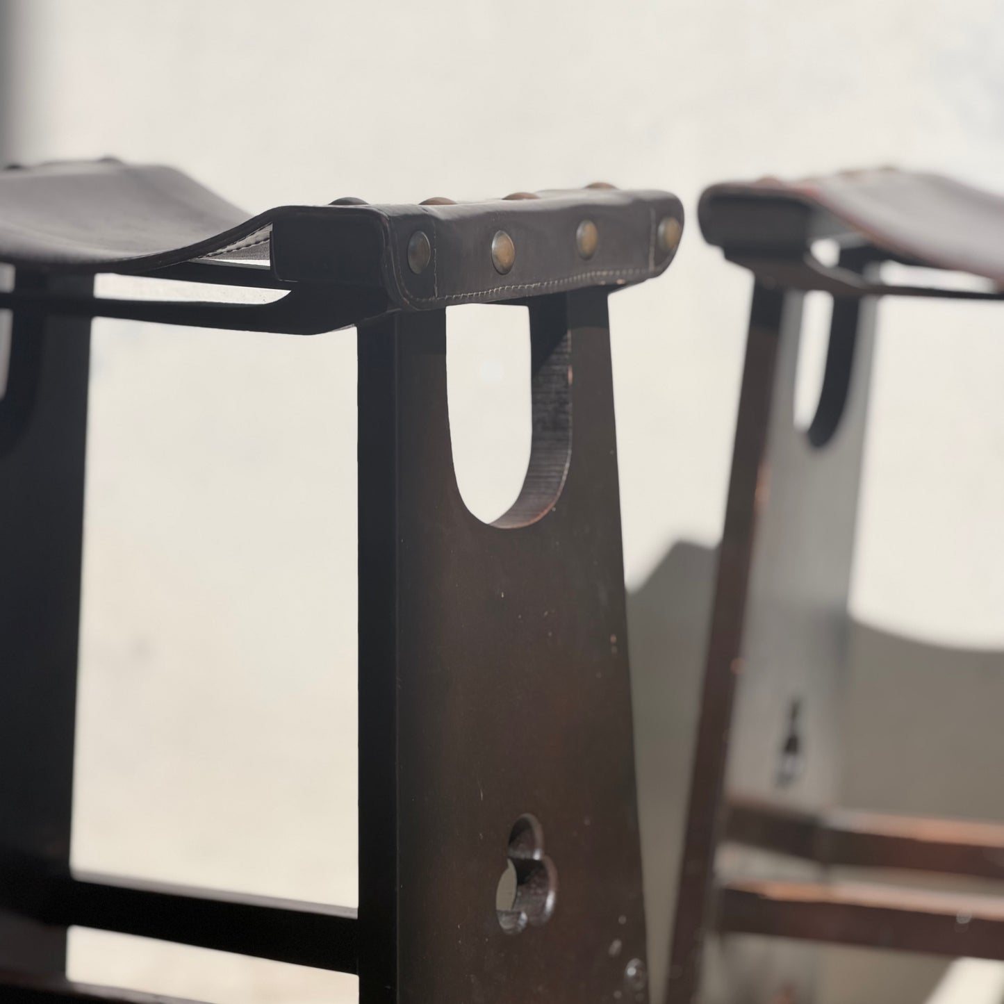 Close-up of a set of three 1970s Australian Mid-Century Modern Spanish Revival Davis Stools (Revive), showcasing dark wood, leather seats with metal rivets, and partial shadows on a plain light background.
