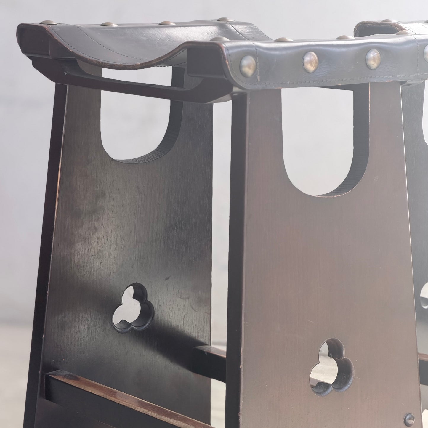 Close-up of two 1970s Australian Mid-Century Modern Spanish Revival Davis Stools (Set of 3, Revive) with black leather seats, metal rivets, and signature trefoil cut-out legs by R.G. Davis Furniture, shown against a softly lit background.