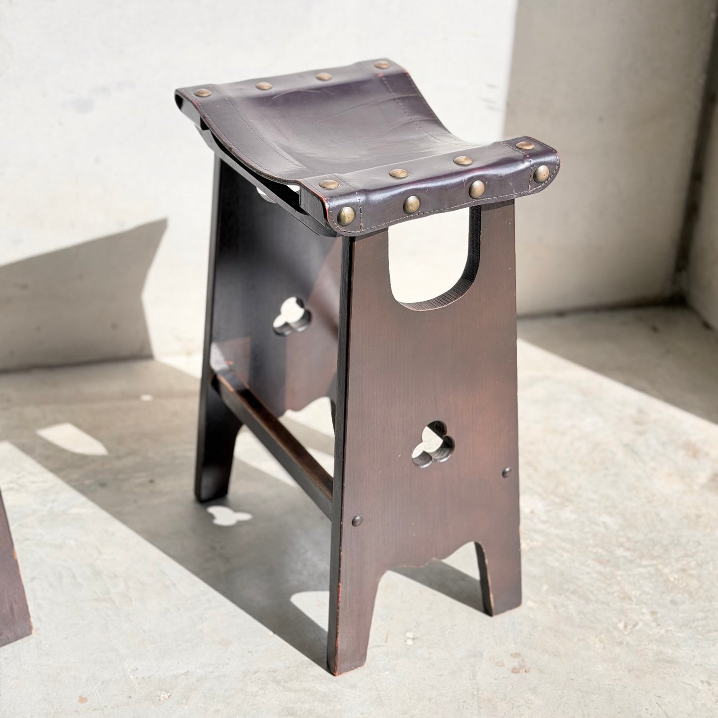 A set of three 1970s Australian Mid-Century Modern Spanish Revival Davis Stools (Revive) features curved leather seats with brass studs and dark wooden frames, shown on a sunlit concrete floor enhanced by light and shadow.