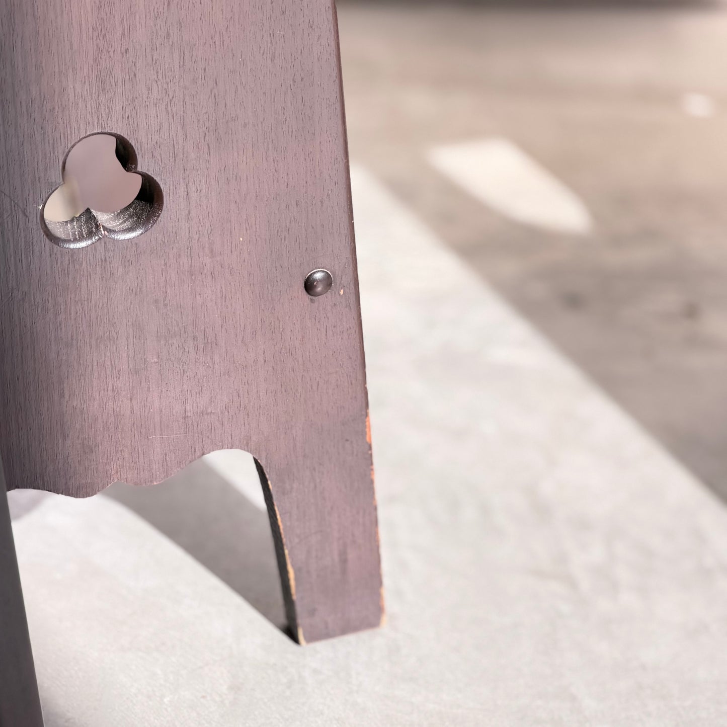 A close-up of a leg from the 1970s Australian Mid-Century Modern Spanish Revival Davis Stools – Set of 3 (Revive), featuring a clover-shaped cutout and metal screw, standing on concrete with sunlight and soft shadows.