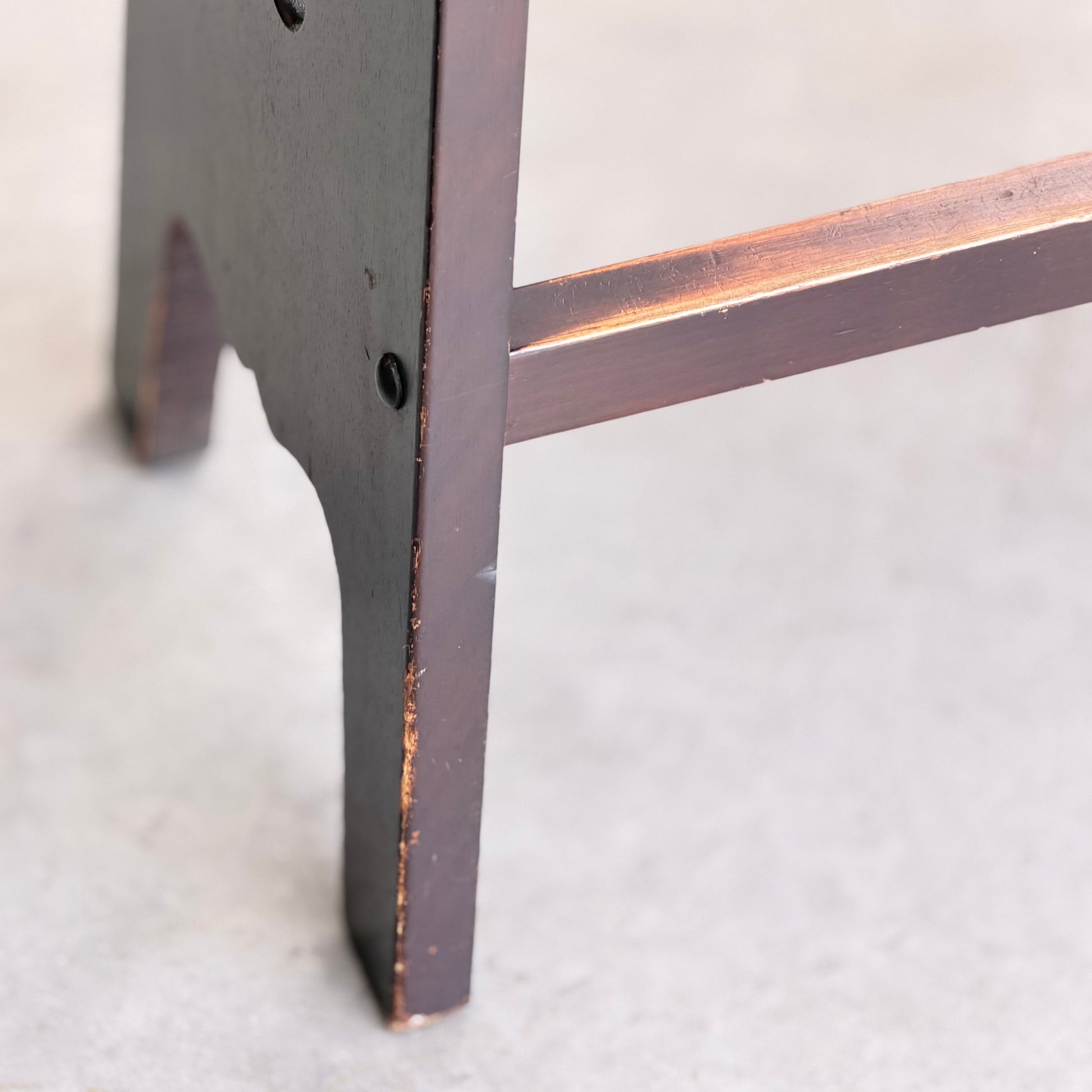 Close-up of the leg and support bar of a 1970s Australian Mid-Century Modern Spanish Revival Davis Stool from the Revive set of 3, showing visible wear on the dark wood finish, set against a light-colored floor.