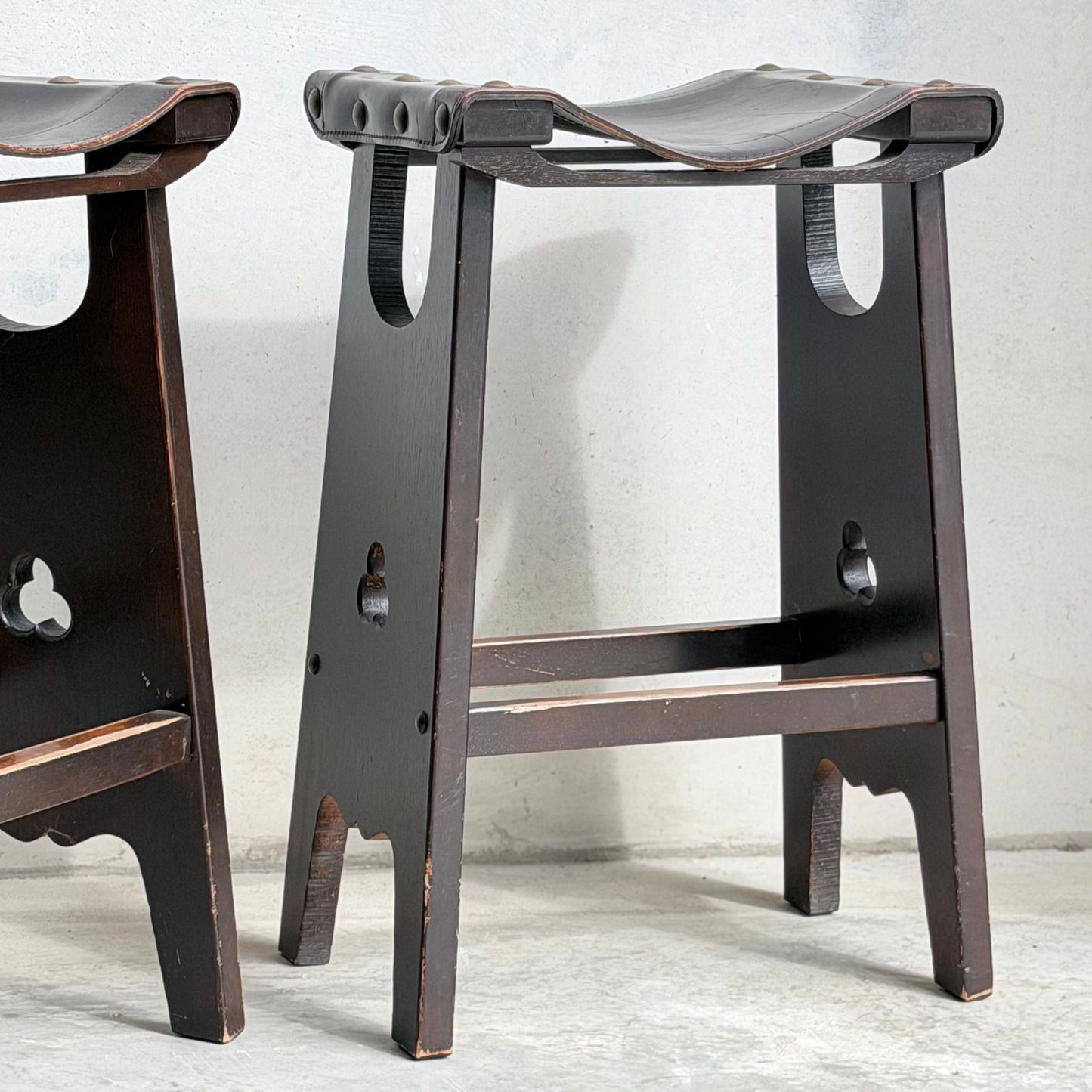 A close-up of the 1970s Australian Mid-Century Modern Spanish Revival Davis Stools – Set of 3 (Revive), showing curved seats and leg cutouts, set on a light concrete floor against a textured white wall.