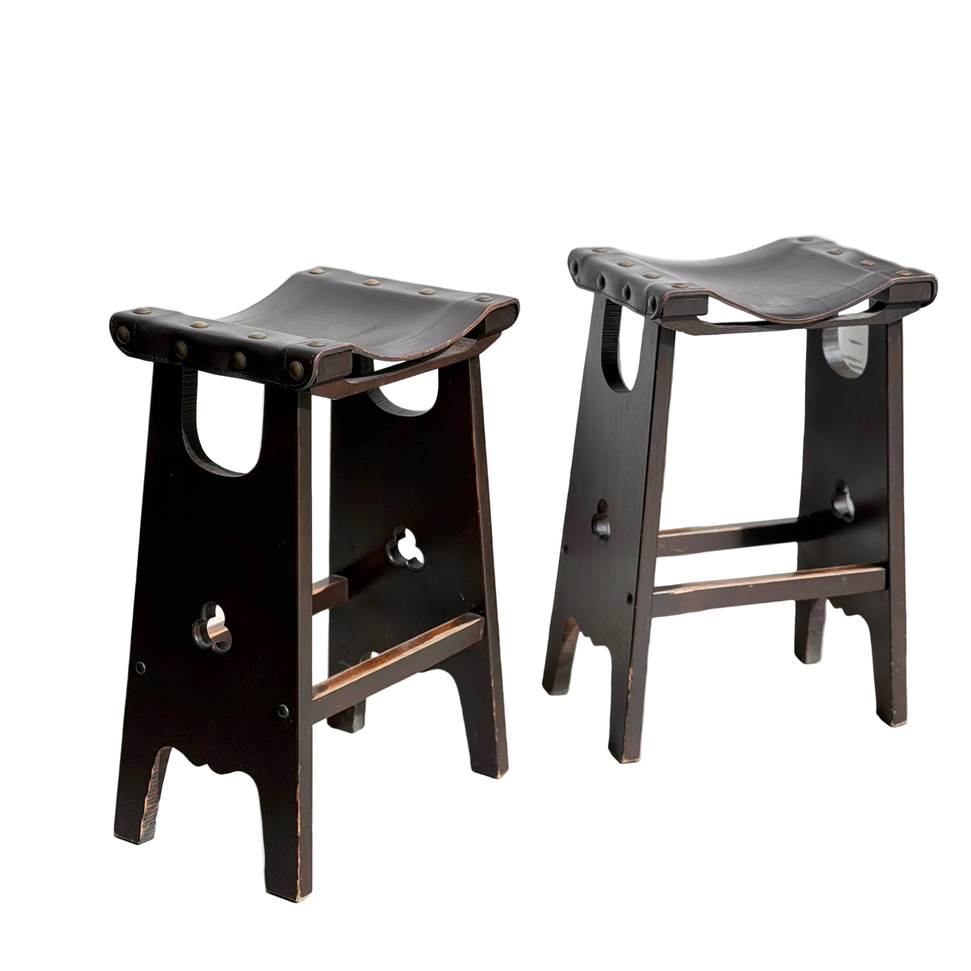 A set of three 1970s Australian Mid-Century Modern Spanish Revival Davis Stools (Revive) with dark wood, curved tops, leather seats, cut-out details, and metal rivets sits on a sunlit concrete floor against a light wall.