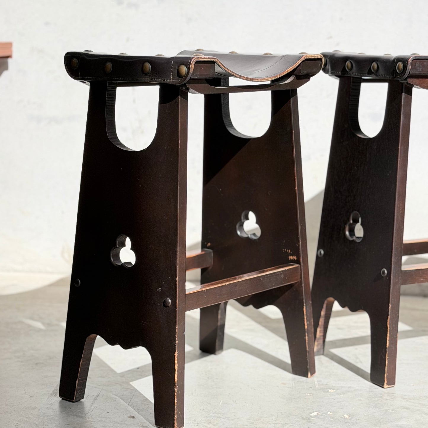 Three 1970s Australian Mid-Century Modern Spanish Revival Davis stools from Revive, with dark wood and leather, stand on a sunlit floor by a pale wall, reflecting classic craftsmanship.