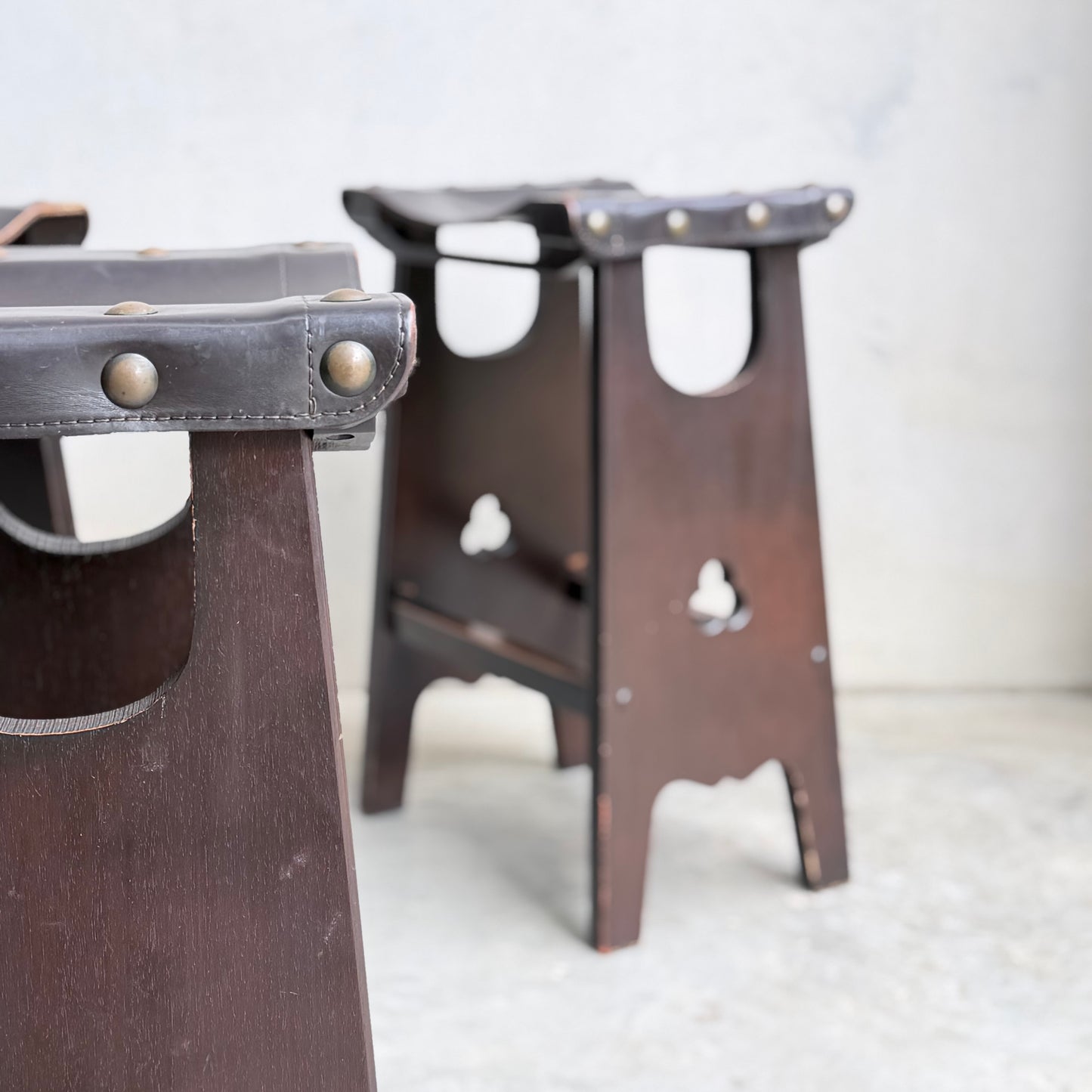 Two dark wooden 1970s Australian Mid-Century Modern Spanish Revival Davis Stools from Revive, featuring leather seats with metal studs, rest on a light concrete floor against a pale wall—one in the foreground, one further back.