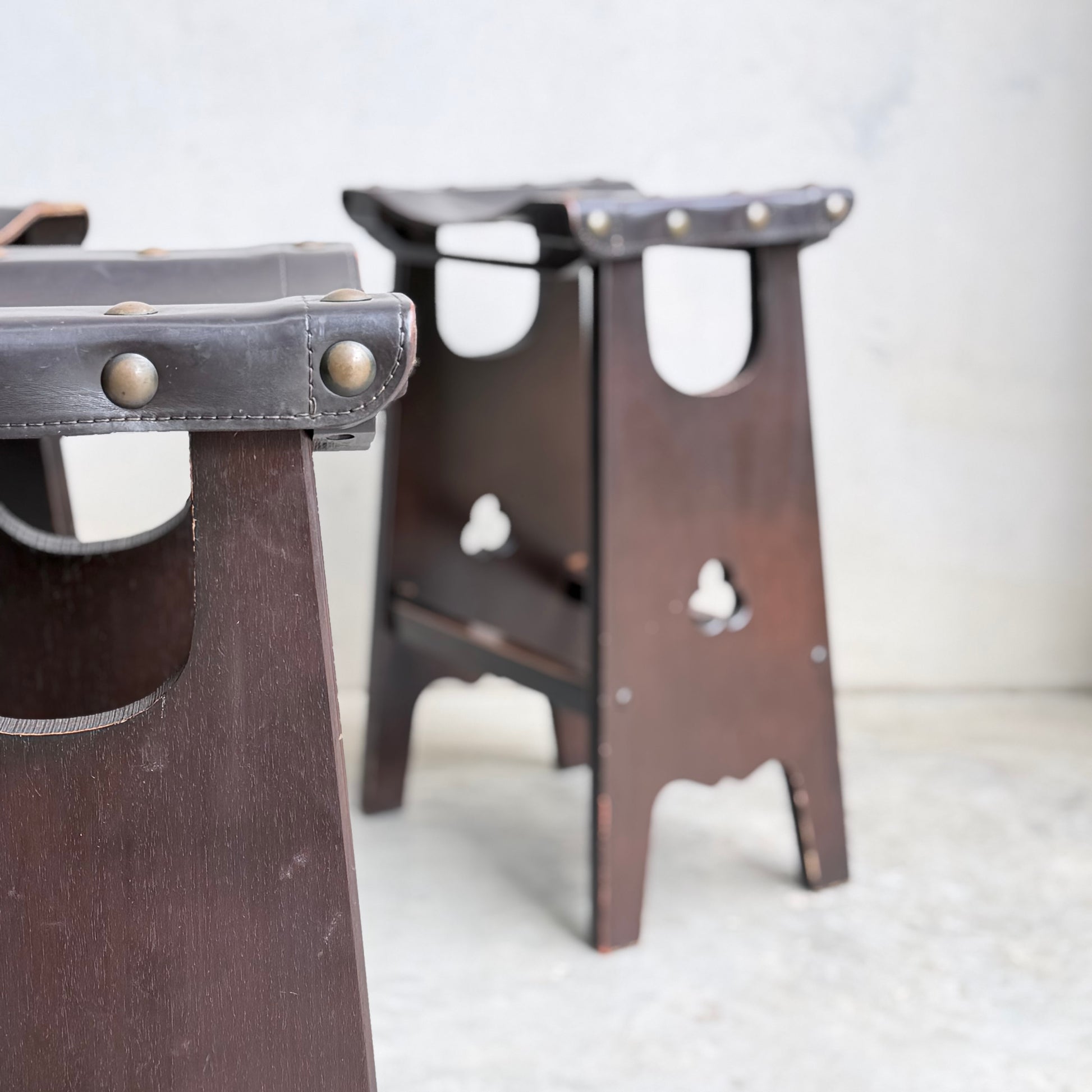 Two dark wooden 1970s Australian Mid-Century Modern Spanish Revival Davis Stools from Revive, featuring leather seats with metal studs, rest on a light concrete floor against a pale wall—one in the foreground, one further back.