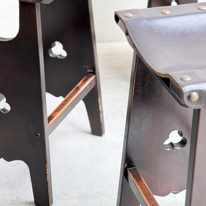 Close-up of two 1970s Australian Mid-Century Modern Spanish Revival Davis Stools (Set of 3), featuring leather seats with cut-out patterns, metal rivets, and dark wood frames showing scuffs and wear, set on a light-colored floor.