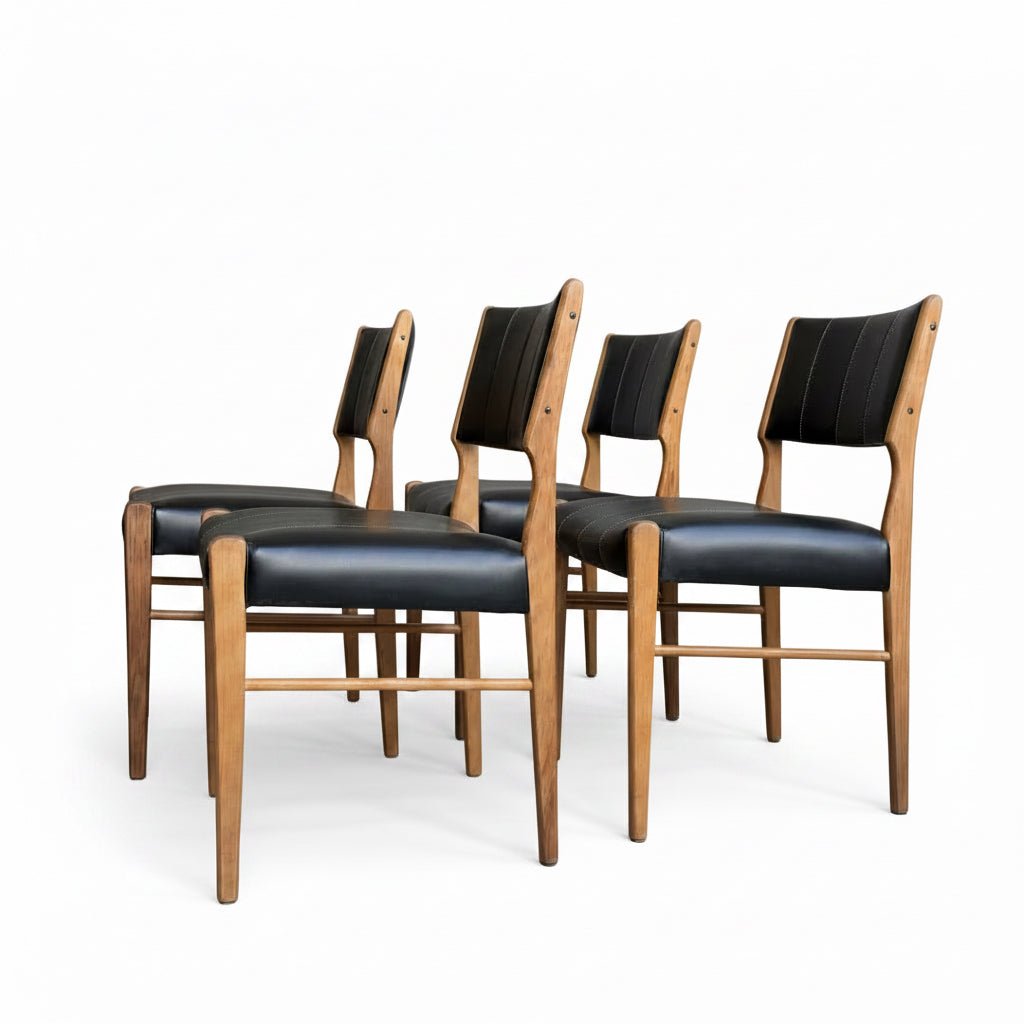 A set of four Vintage TH Brown Blackwood Dining Chairs – Set of 4 (Revive), featuring black upholstered seats and backs, are lined up on a smooth grey floor, with part of a wooden table visible to the left.