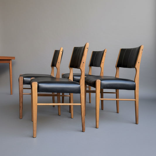 A set of four Vintage TH Brown Blackwood Dining Chairs – Set of 4 (Revive), featuring black upholstered seats and backs, are lined up on a smooth grey floor, with part of a wooden table visible to the left.