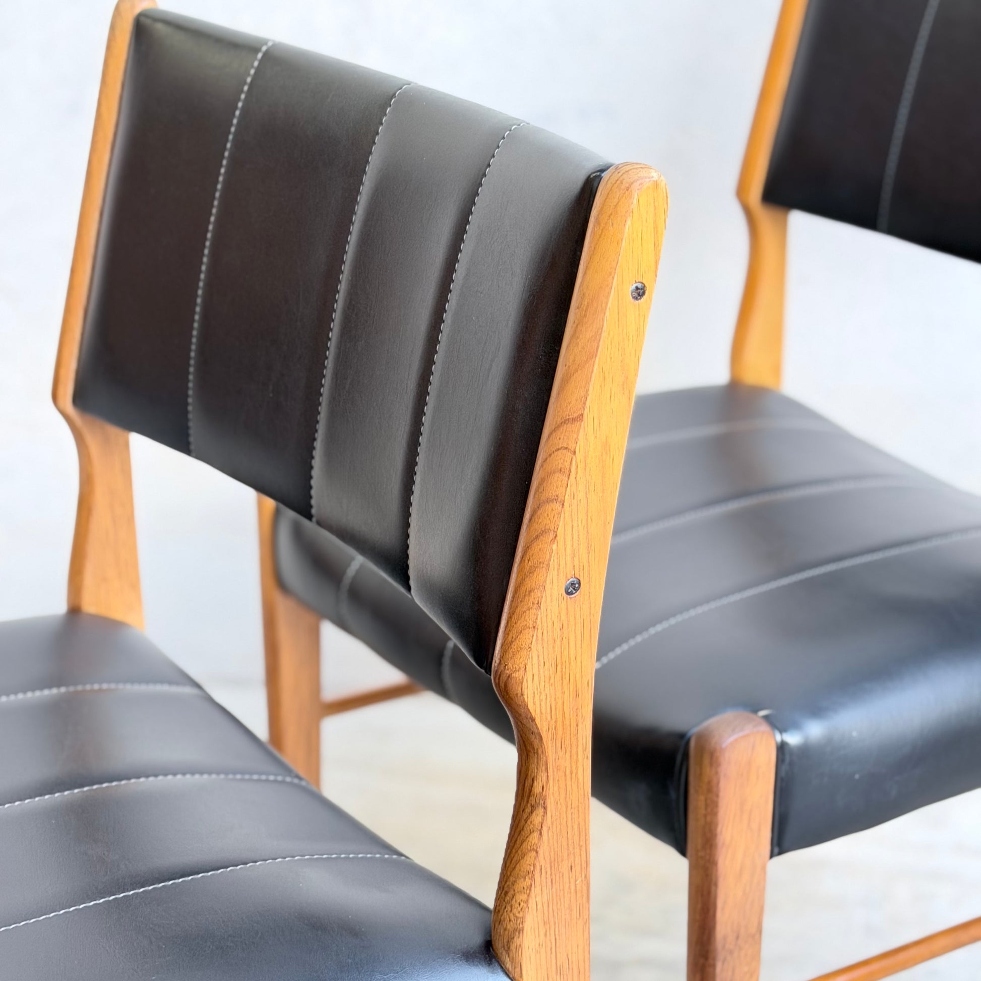 A set of four Vintage TH Brown Blackwood Dining Chairs (Revive) featuring black leather cushions and vertical stitching are arranged on a light floor, highlighting classic mid-century modern style.