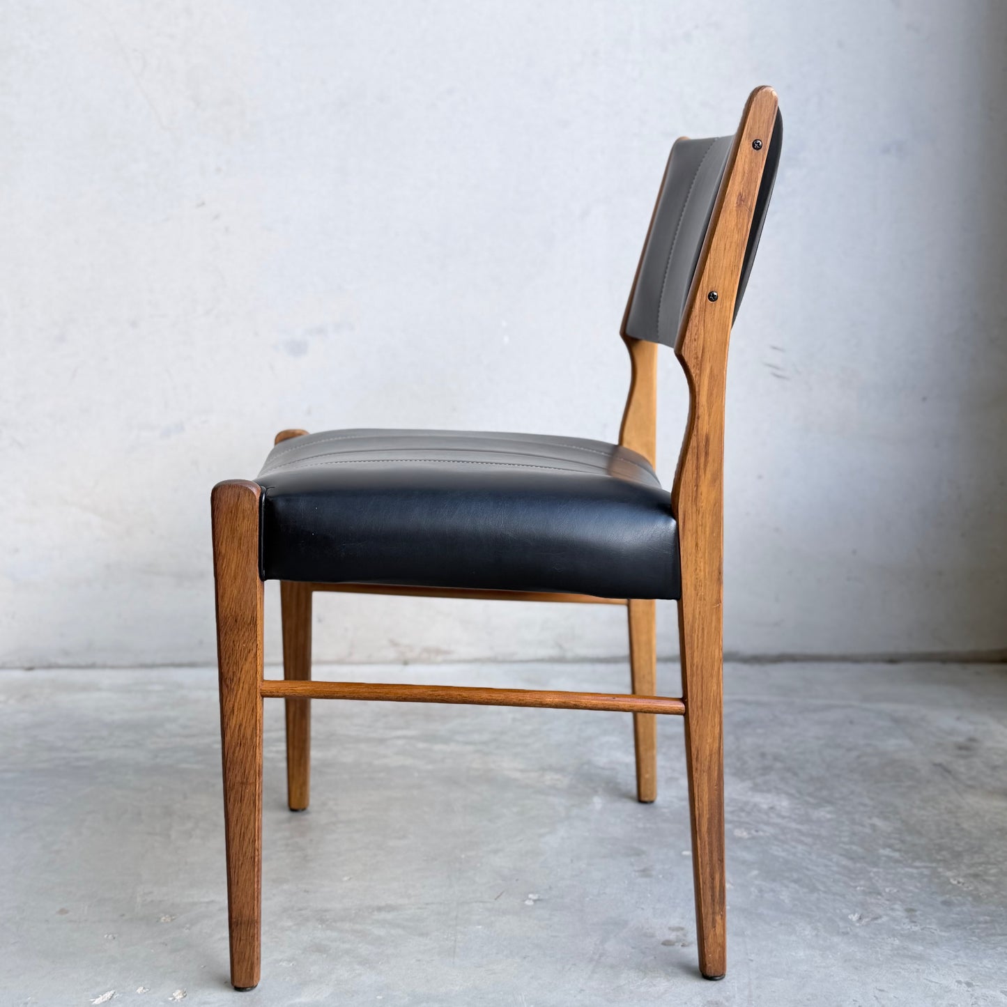 A set of four Vintage TH Brown Blackwood Dining Chairs (Revive) featuring black leather seats and backrests, mid-century modern style, wooden frames, viewed from the side against a light wall and concrete floor.