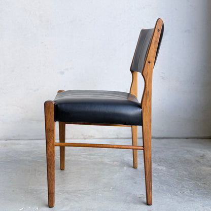 A set of four Vintage TH Brown Blackwood Dining Chairs (Revive) featuring black leather seats and backrests, mid-century modern style, wooden frames, viewed from the side against a light wall and concrete floor.