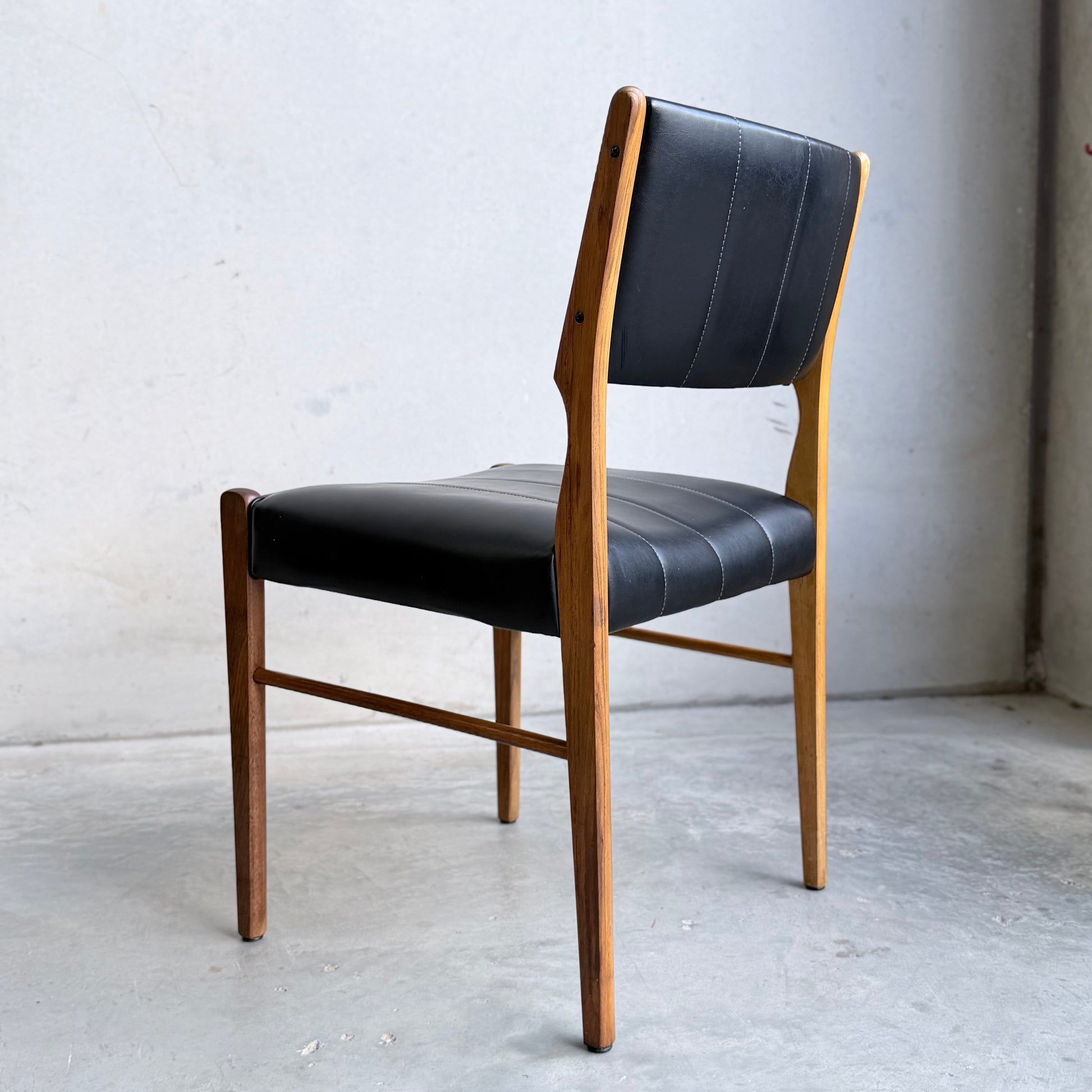 A set of four Vintage TH Brown Blackwood Dining Chairs (Revive), featuring black cushioned seats, wooden frames, and tapered legs, is arranged on a concrete floor against a plain light-colored wall.