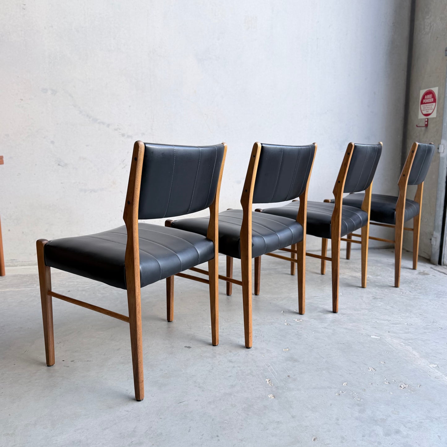 A set of four Vintage TH Brown Blackwood Dining Chairs – Revive edition, with black padded seats and backs, are arranged in a row on a light concrete floor against a plain wall, shown facing away from the camera.