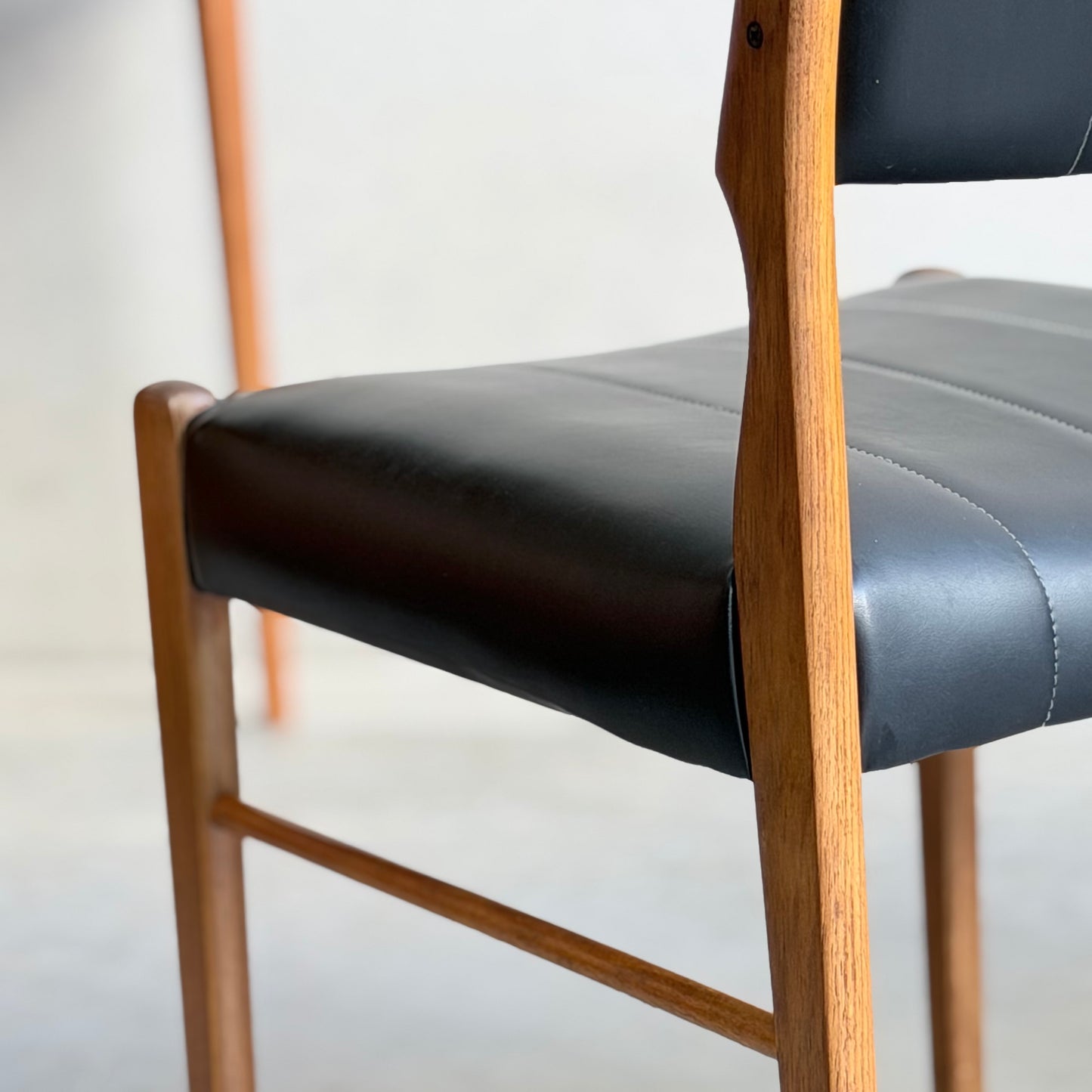 Close-up of a Vintage TH Brown Blackwood Dining Chair from the Revive set of 4, featuring smooth black leather seat and backrest with a natural wood finish. The softly blurred background adds to the mid-century modern appeal.